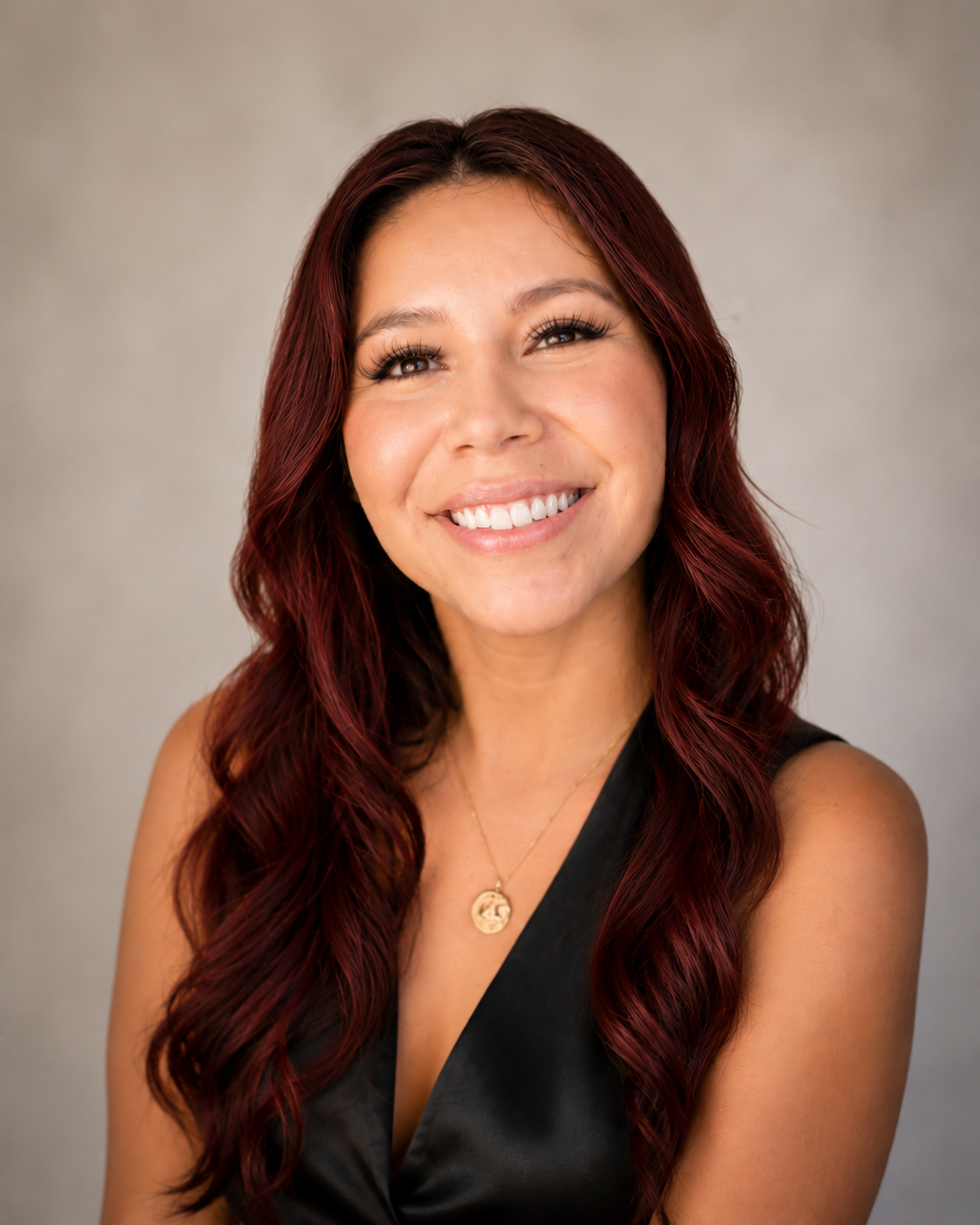 A woman with long wavy red hair smiling, wearing a black sleeveless top and a gold necklace with a circular pendant.
