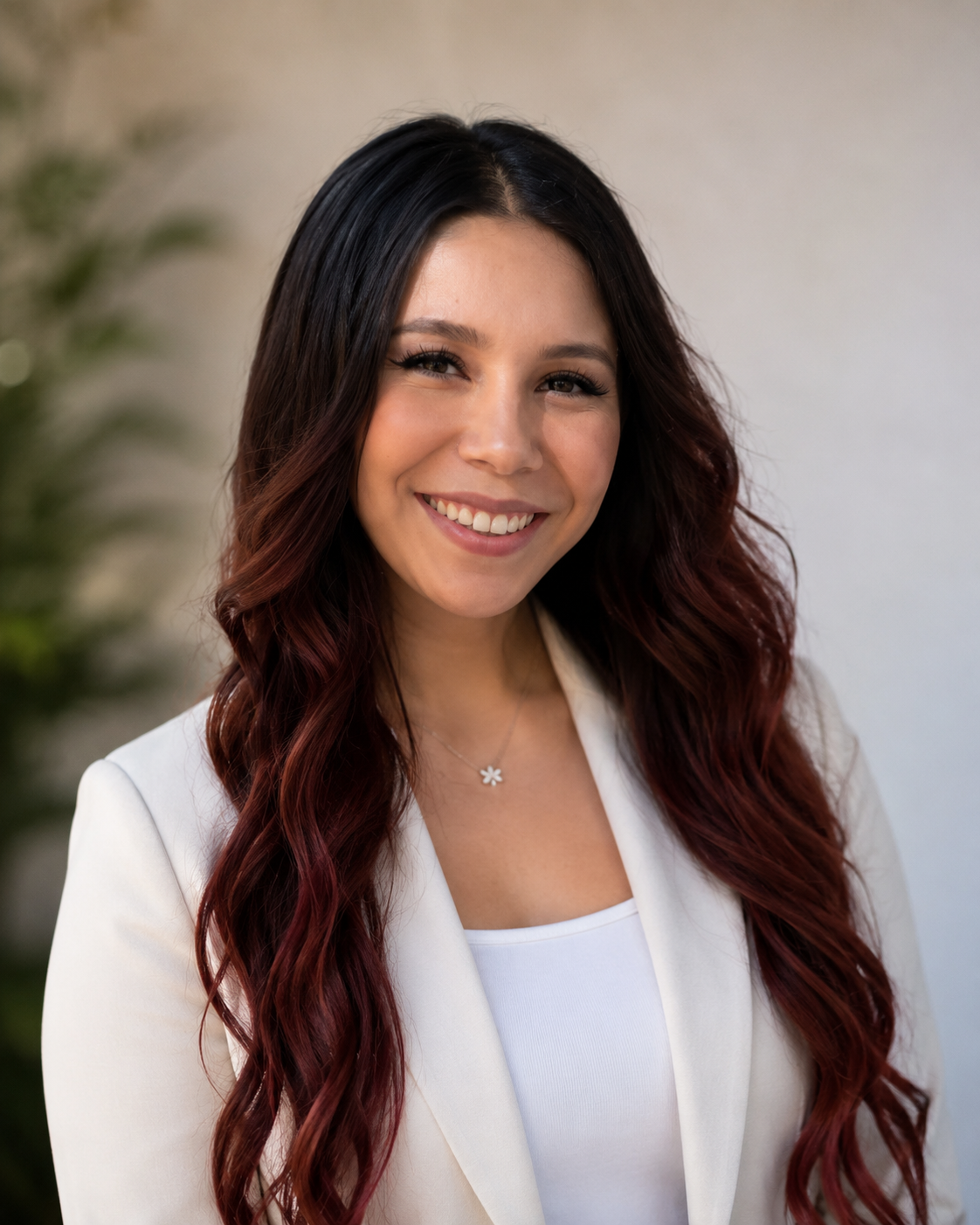 A woman with long wavy dark hair with red highlights, smiling, wearing a white blazer and a daisy pendant necklace.