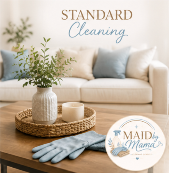 Living room with a wooden coffee table holding a white vase with green leaves, a candle, and blue cleaning gloves, with a beige sofa and pillows in the background. The wall has the words 'STANDARD Cleaning' and a circular logo for 'Maid by Mama Cleaning Services'.