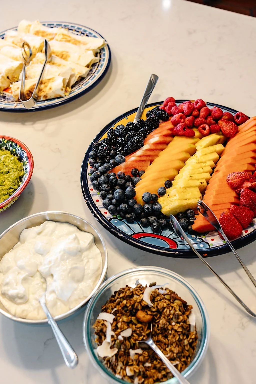 Assorted fresh fruit platter with slices of watermelon, cantaloupe, pineapple, and strawberries, surrounded by bowls of granola, yogurt, guacamole, and scrambled eggs on a breakfast table.