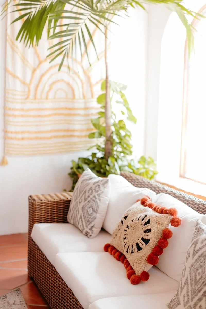 A wicker sofa with white cushions and patterned pillows in a bright room with natural light. A decorative pillow with orange pom-poms and a circular design is on the sofa. Green plants and a wall hanging with a rainbow and stripe pattern are in the b