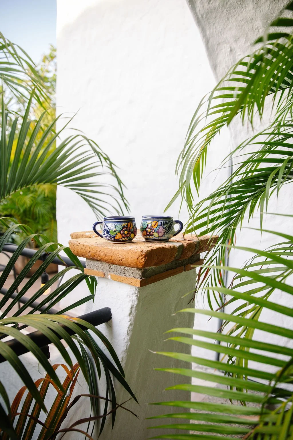 Two colorful ceramic cups with floral patterns sit on a brick and cement ledge, surrounded by lush green palm leaves on a bright outdoor porch.