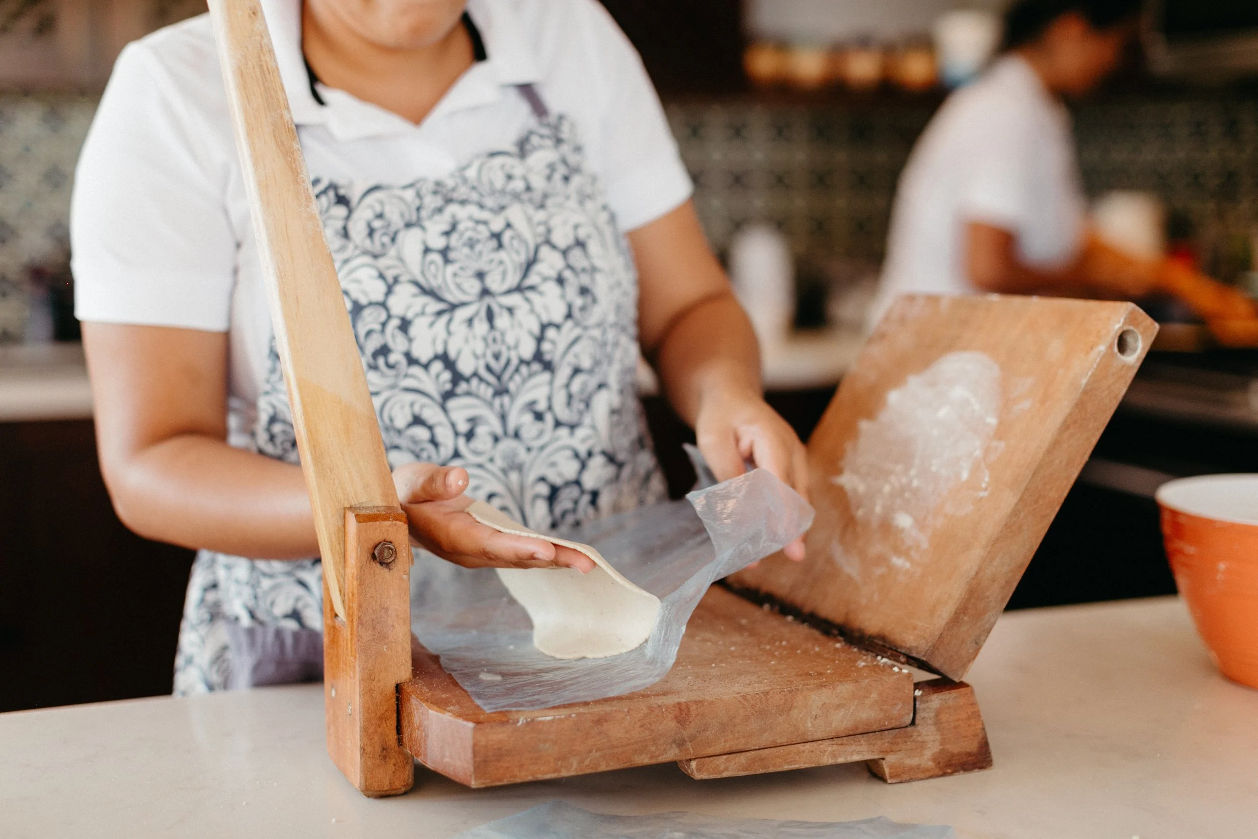 Child holding a piece of paper in front of a wooden press or mold, with another child blurred in the background, possibly involved in a craft or baking activity.