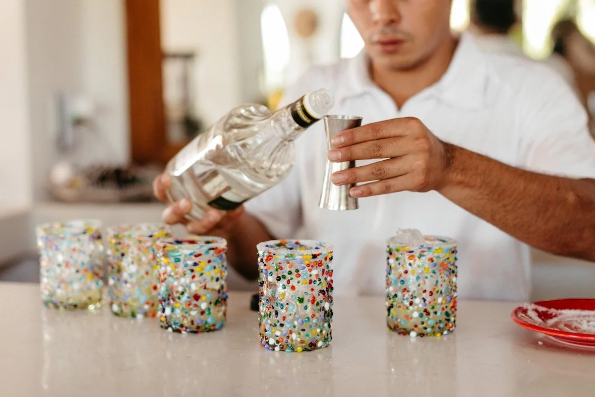 A person pouring liquid from a bottle into a shot glass in front of five decorated cakes covered with colorful sprinkles.
