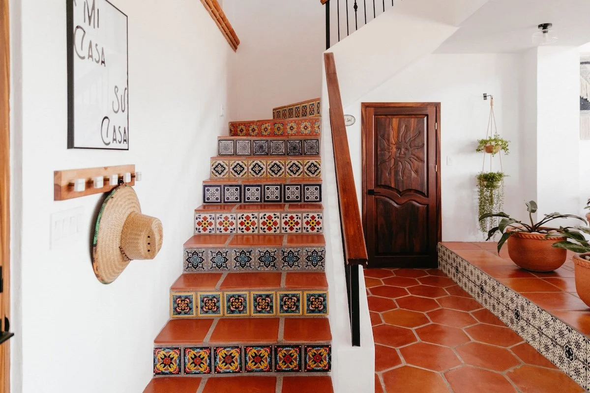 Interior of a house with tiled staircase, wooden door with carved sun design, hanging plants, and terra cotta tiled floors.