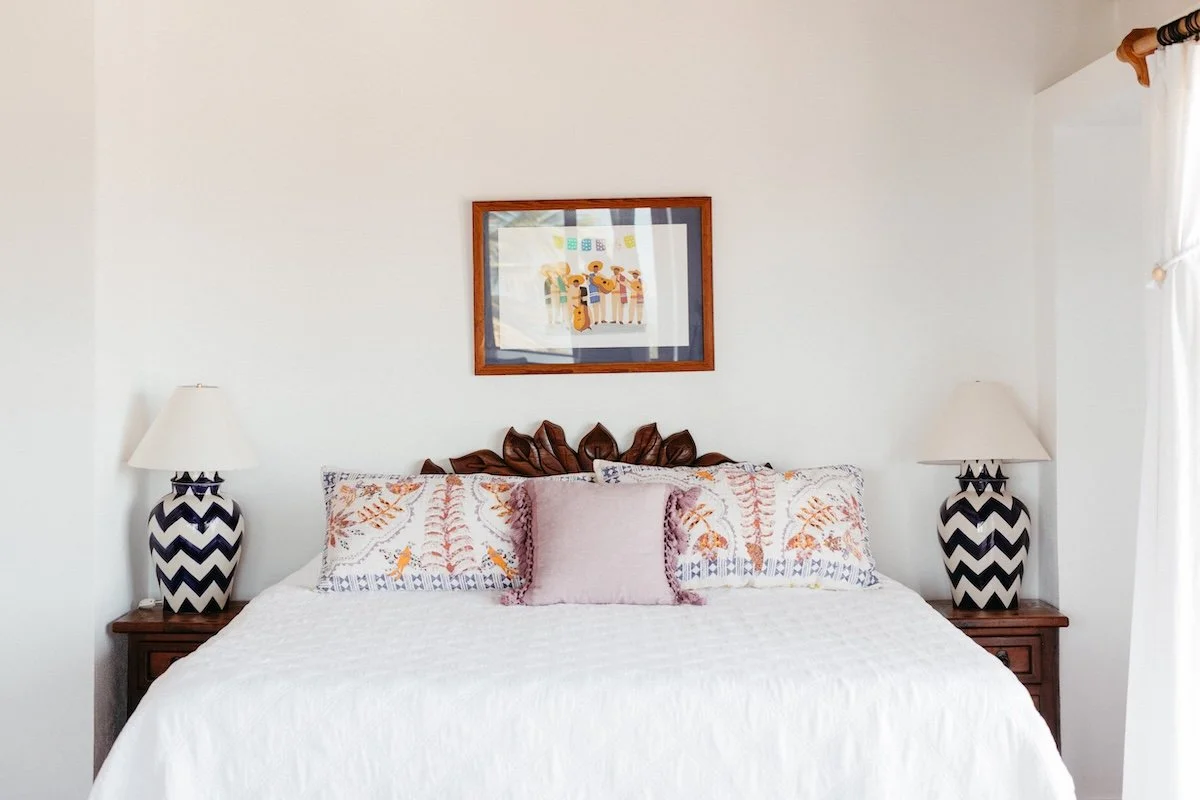 Bedroom with white bedding, decorative pillows, framed picture above bed, and matching nightstands with chevron-patterned lamps on each side.