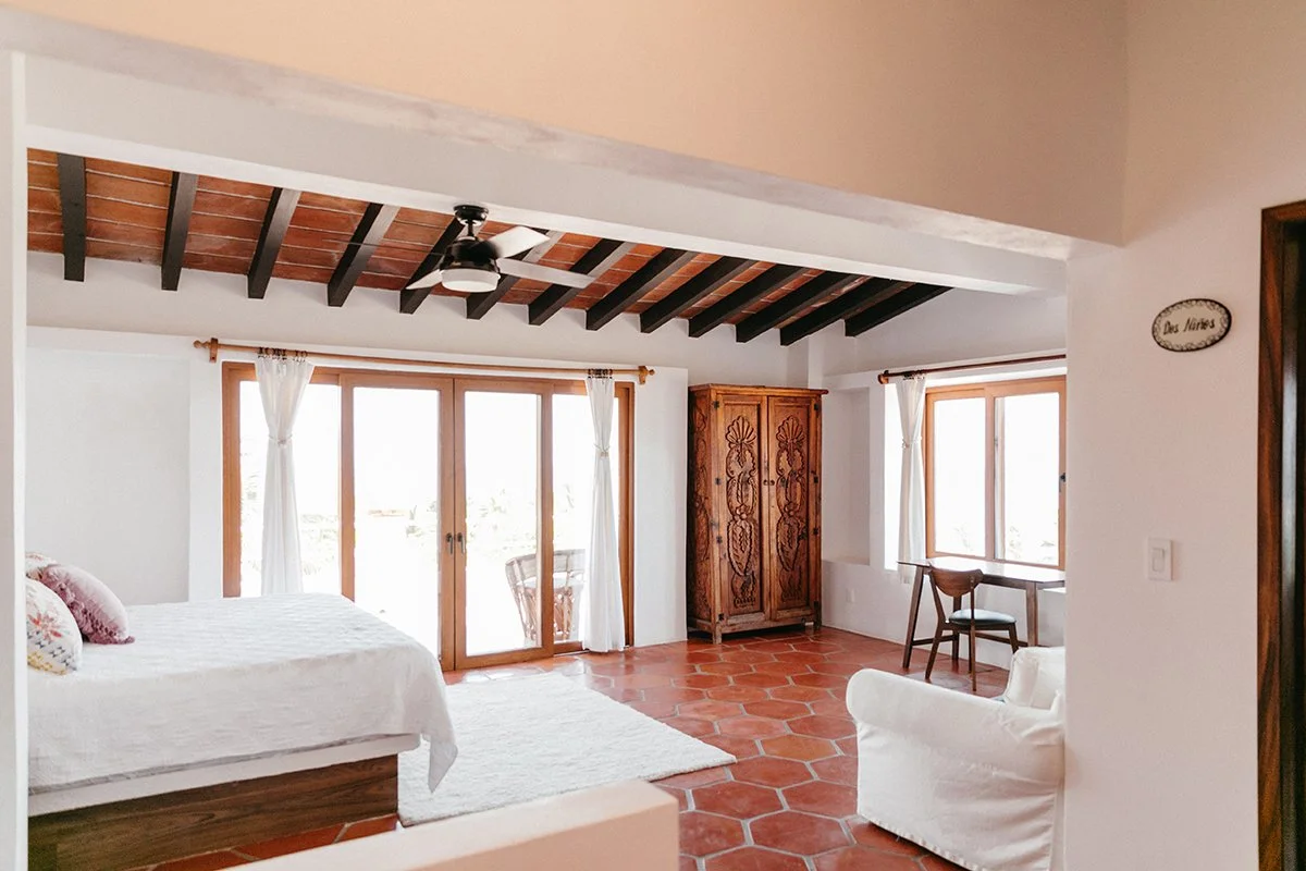 A bright bedroom with wooden ceiling beams, a ceiling fan, large sliding glass doors with white curtains, a bed with white linens and decorative pillows, a wooden armoire, a small desk with a chair near a window, and terracotta tile flooring.