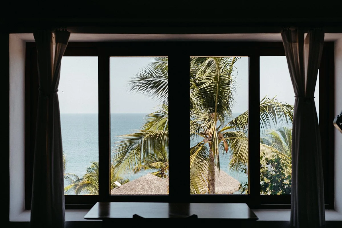 View of palm trees, thatched roofs, and the ocean seen through a window with curtains.
