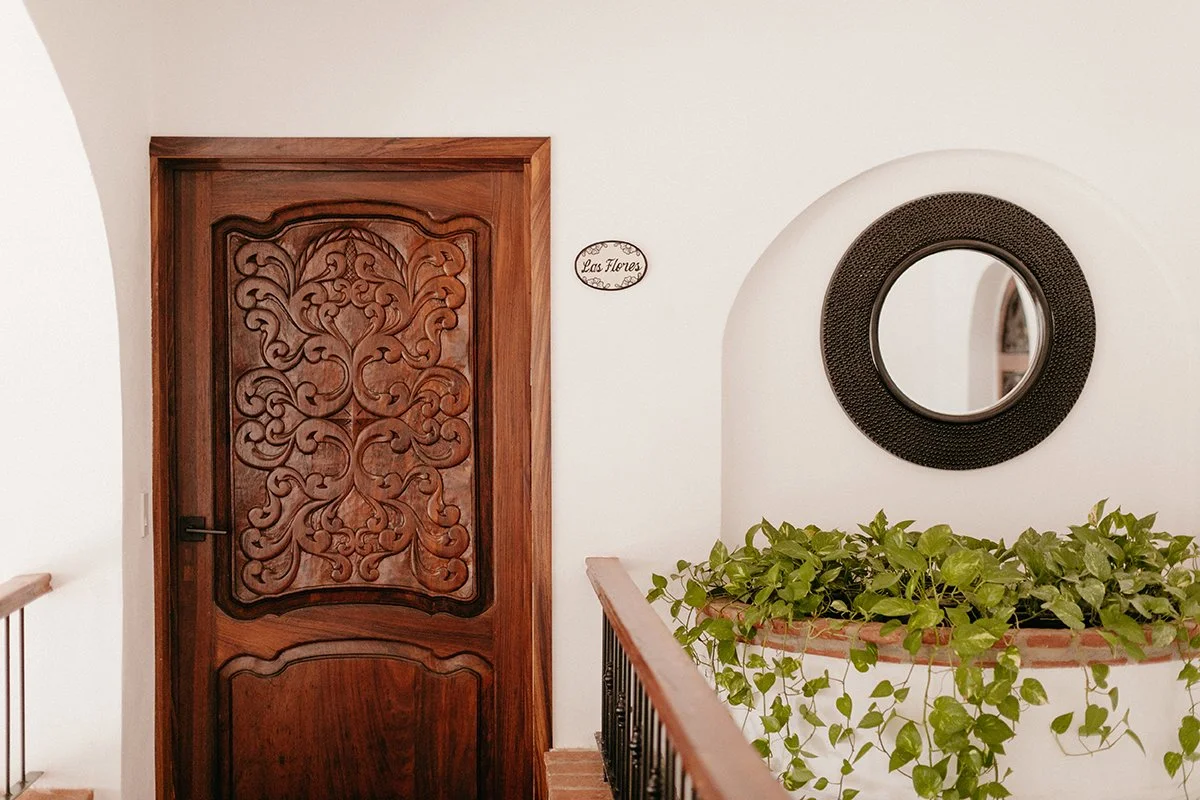 Interior of a house with a carved wooden door, a round mirror on the wall, green plants in a terracotta pot, and a decorative sign that reads 'Las Flores'.