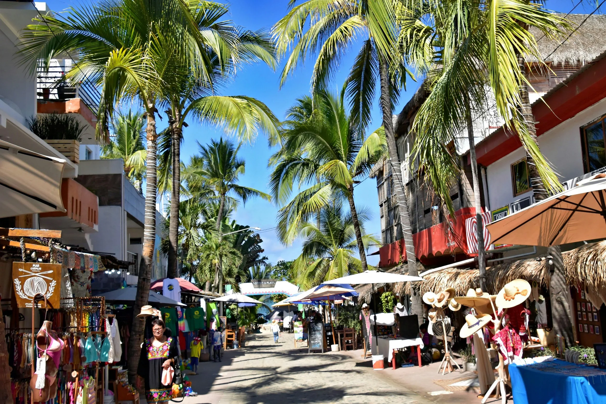 A lively outdoor marketplace lined with colorful stalls selling souvenirs and accessories, with tall palm trees and bright blue sky overhead. There are people walking, shopping, and browsing under umbrellas and shaded areas.