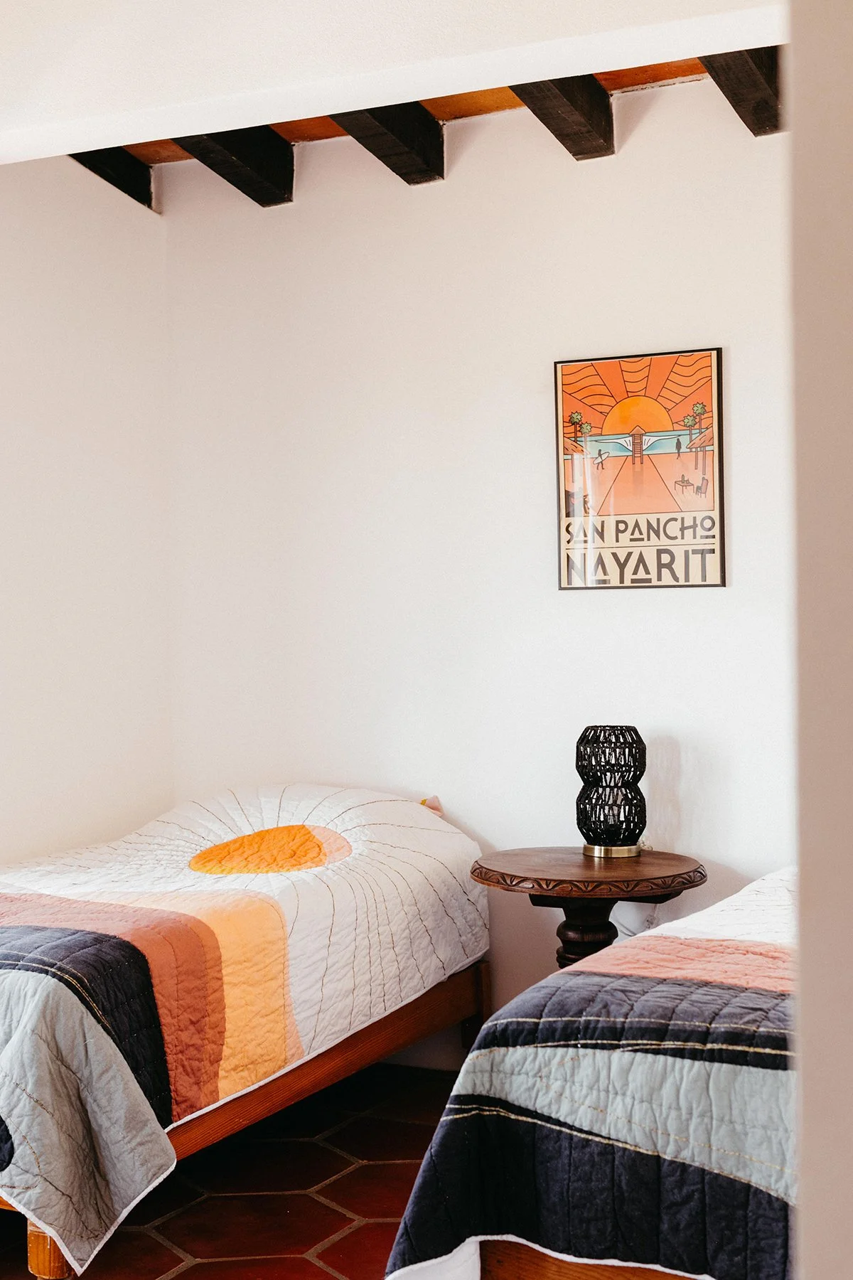 A bedroom with two beds covered with patterned quilts, a small round wooden table with a black woven lamp, and a colorful framed poster of San Pancho Nayarit on the wall.