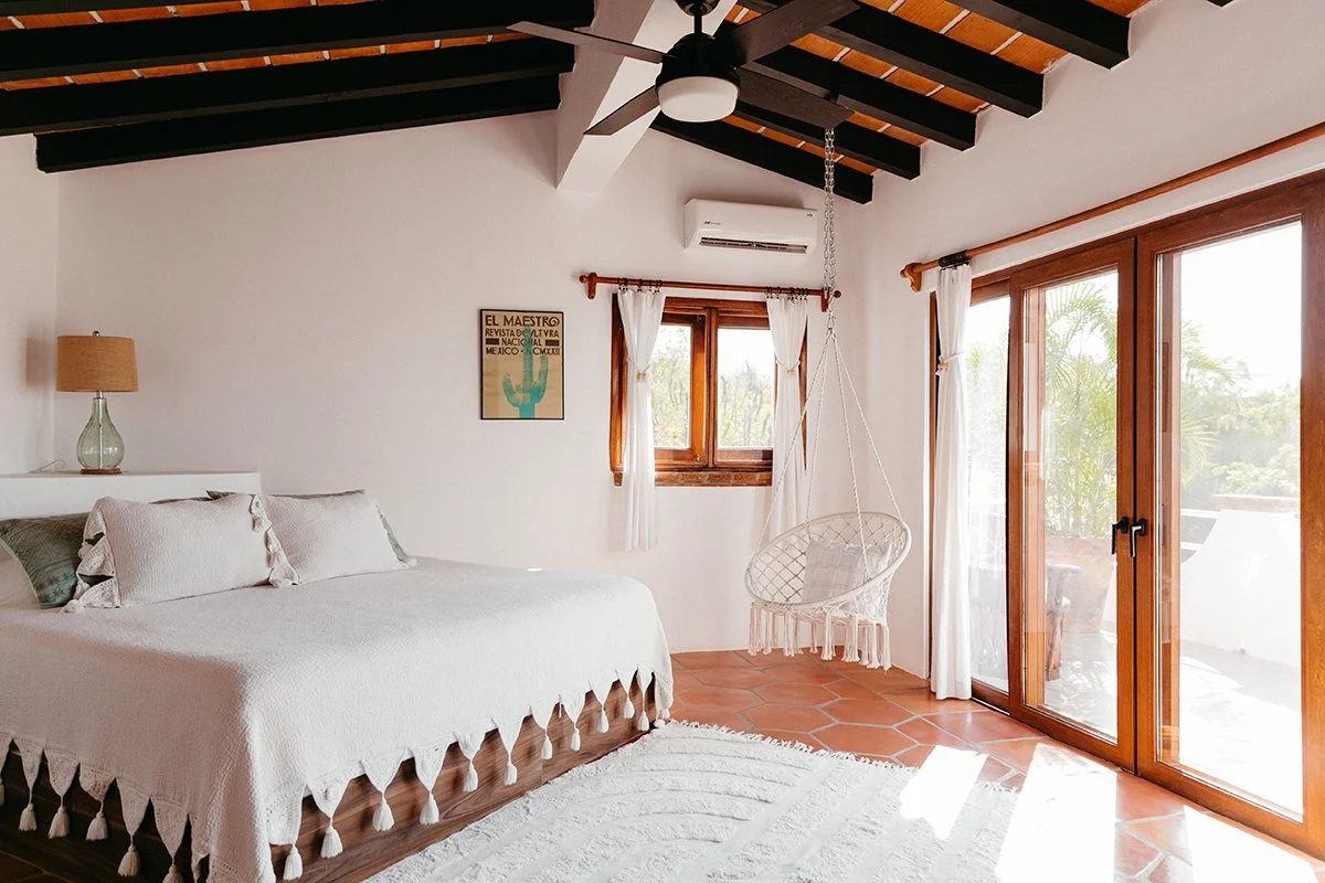 Bright bedroom with white walls, wooden ceiling beams, large sliding glass door with white curtains, a bed with a white bedspread and pillows, a wall lamp on a bedside table, a framed poster on the wall, a hanging wicker chair, and terracotta floor t