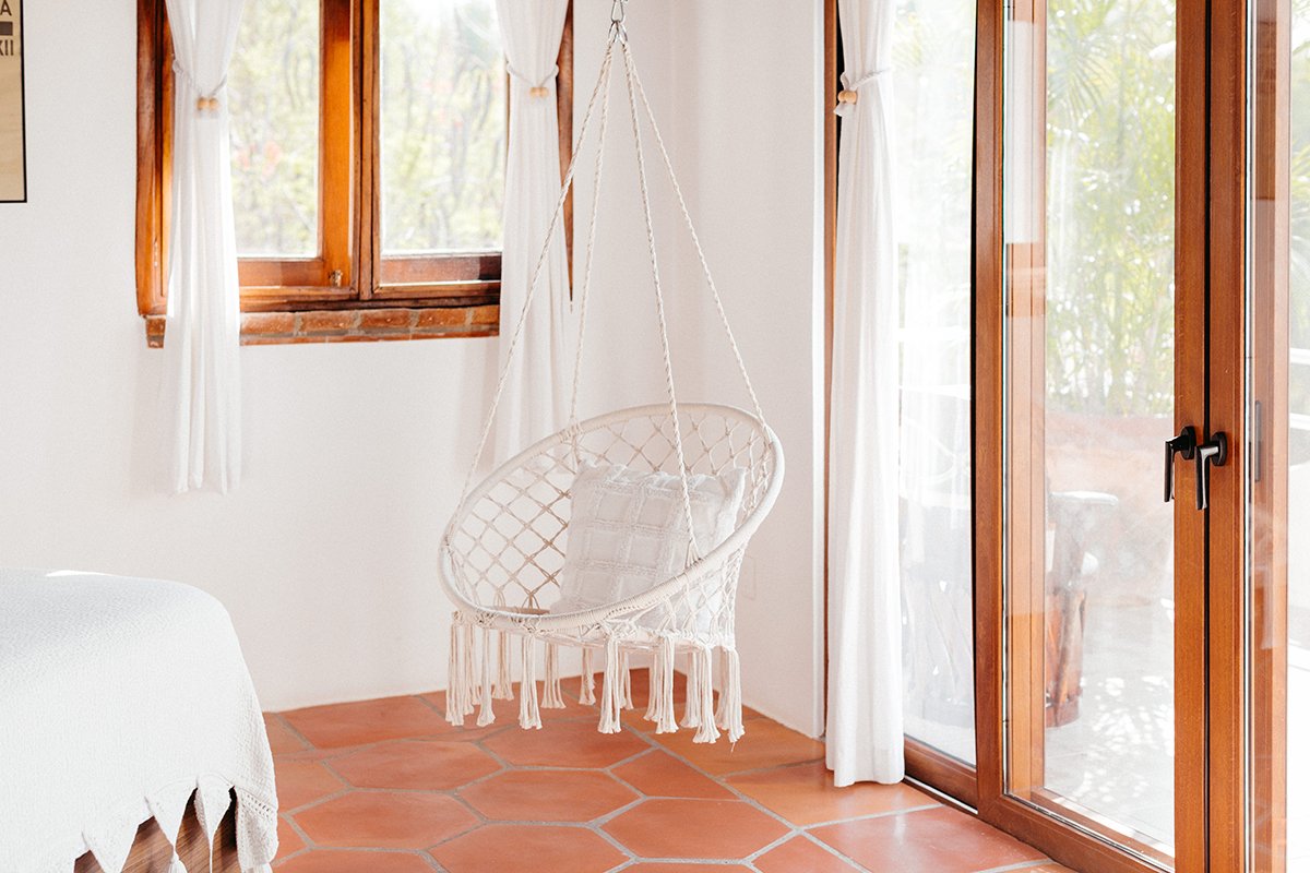 A cozy sunlit corner of a room with a hanging woven chair with a pillow, terracotta tile floor, white curtains, and wooden-framed windows and door.