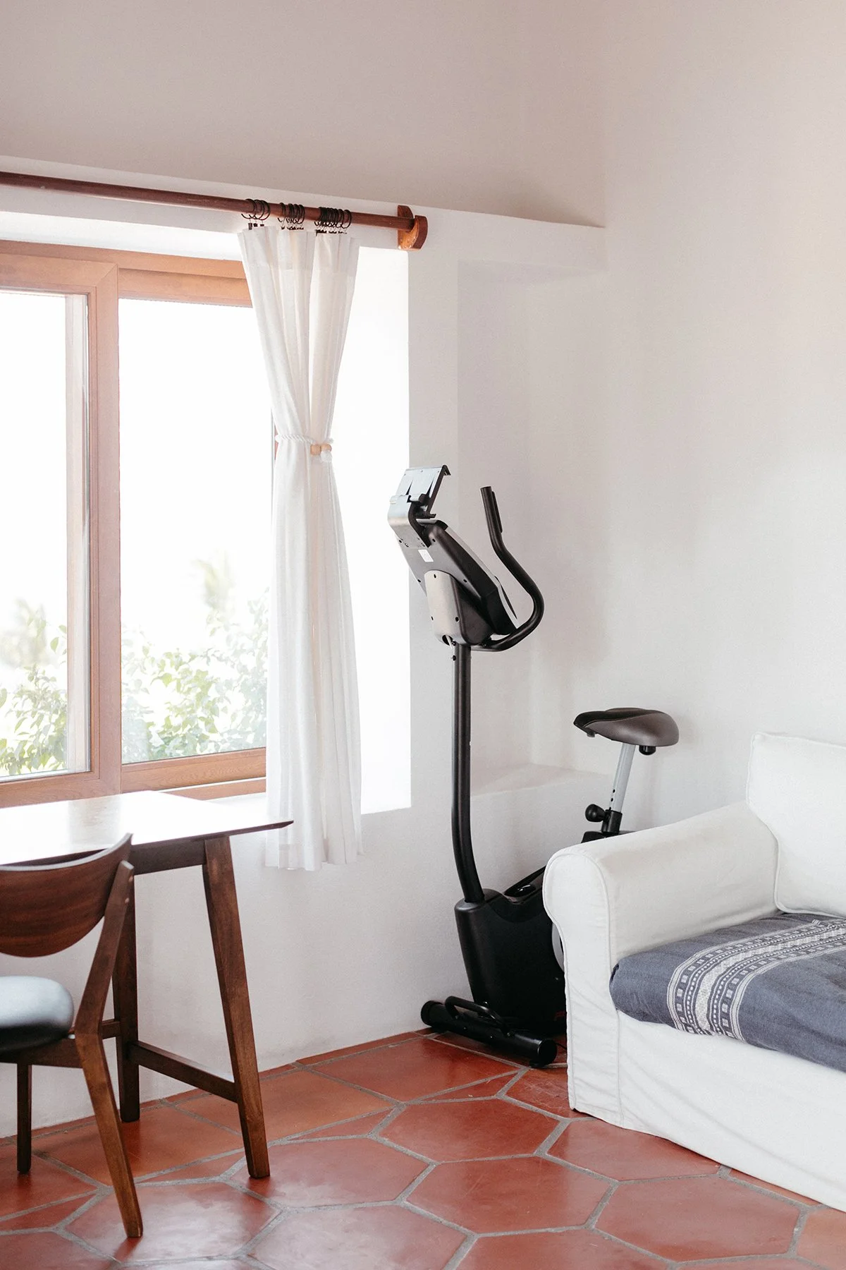 A room with terracotta tiled floors, white walls, and a large window with white curtains, a wooden curtain rod, an exercise bike near a white sofa, and a wooden dining table with chairs.