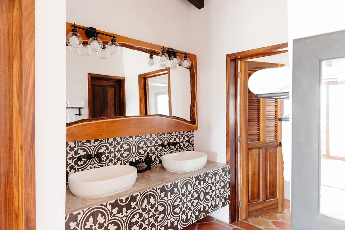 Double bathroom vanity with two white vessel sinks, a wooden framed mirror, and black wall-mounted fixtures, featuring a decorative black and white patterned tile backsplash, wooden door, and wooden window shutters.