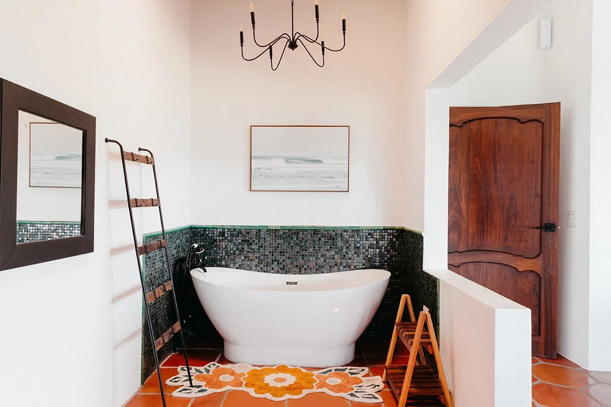 Bathroom with a white freestanding bathtub, a black mosaic tiled wall, a wooden ladder, a framed mirror, a painting of waves, a chandelier, a wooden door, and a colorful floral rug.