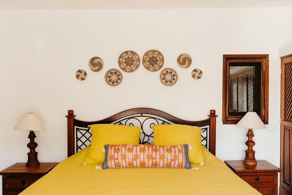 A bedroom with a wooden bed frame, yellow bedding, and decorative pillows. Two wooden nightstands with lamps flank the bed. Woven wall art decorating the wall above the bed and a mirror on the right side of the wall.