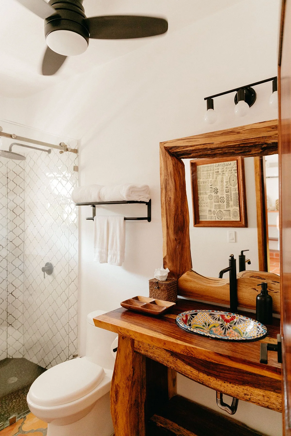 A bathroom with a wooden vanity featuring a colorful patterned sink, a mirror with a wood frame, a black faucet, black soap dispenser, a wall-mounted shelf with a tissue box, a towel rack with two white towels, a shower area with patterned tiles, a c