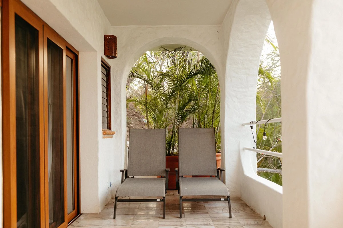 Two lounge chairs on a screened balcony with potted plants and view of trees outside.