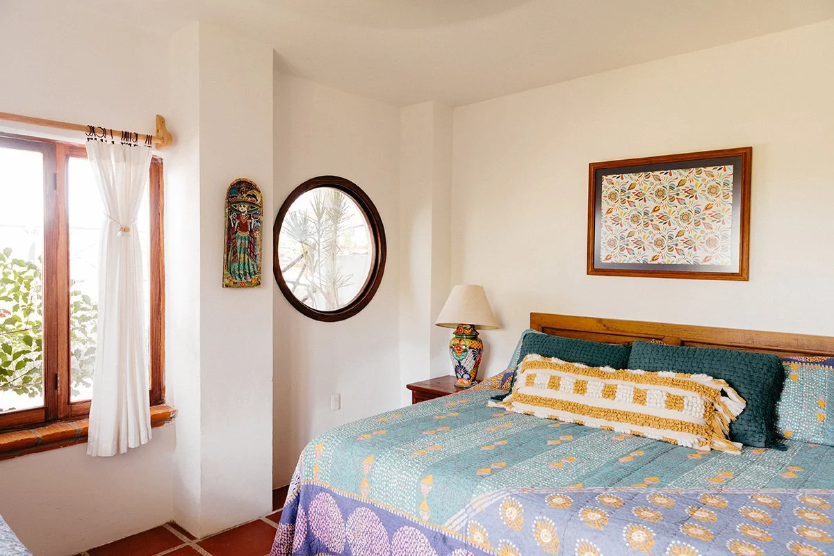 A bedroom with a bed, colorful quilt, decorative pillows, a wooden side table with a floral lamp, a framed abstract artwork above the bed, a window with white curtains, and a small circular mirror on the wall.