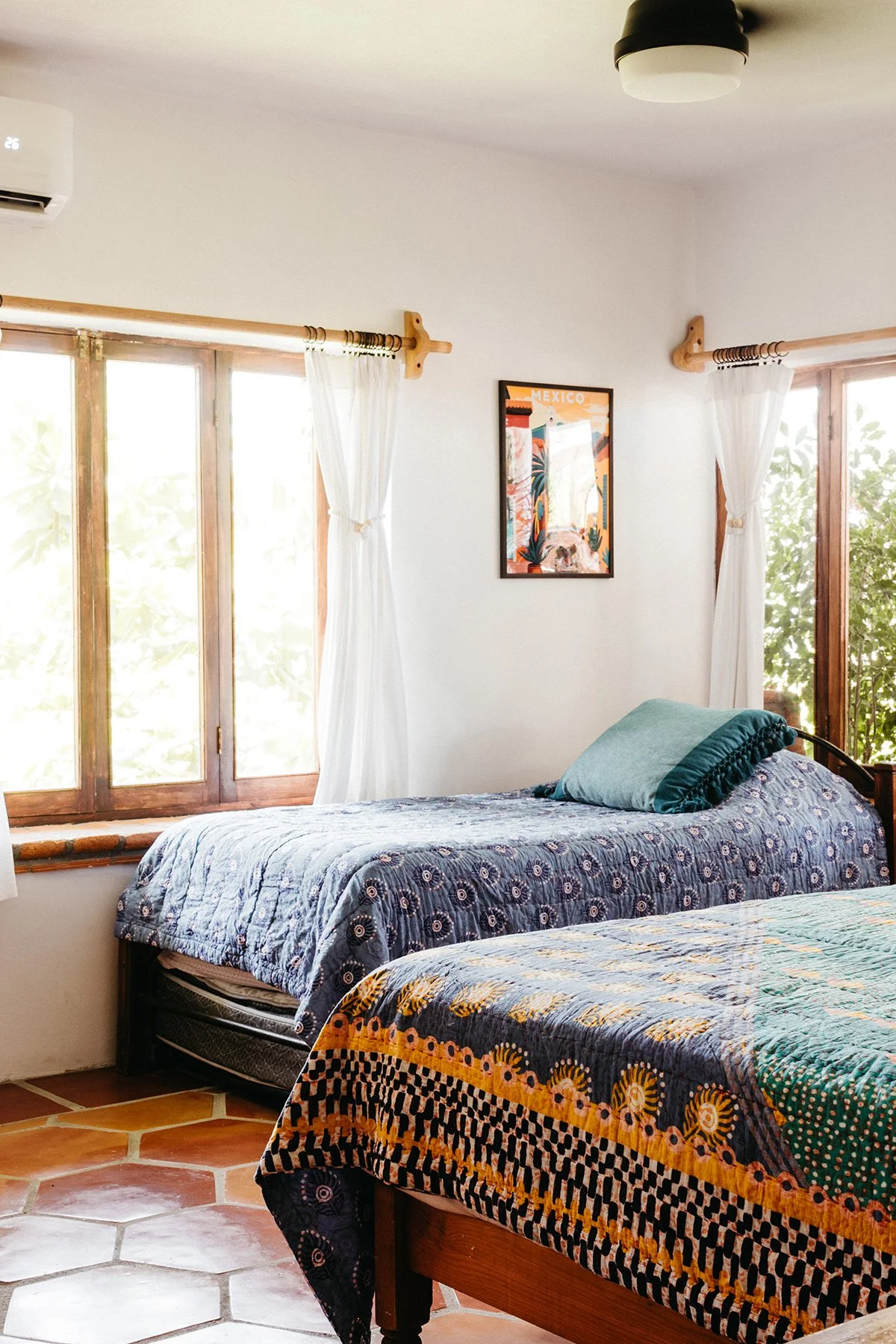 A bedroom with two beds, each with patterned bedspreads and pillows. The room has large wooden-framed windows with white curtains, and a framed colorful travel poster on the wall.