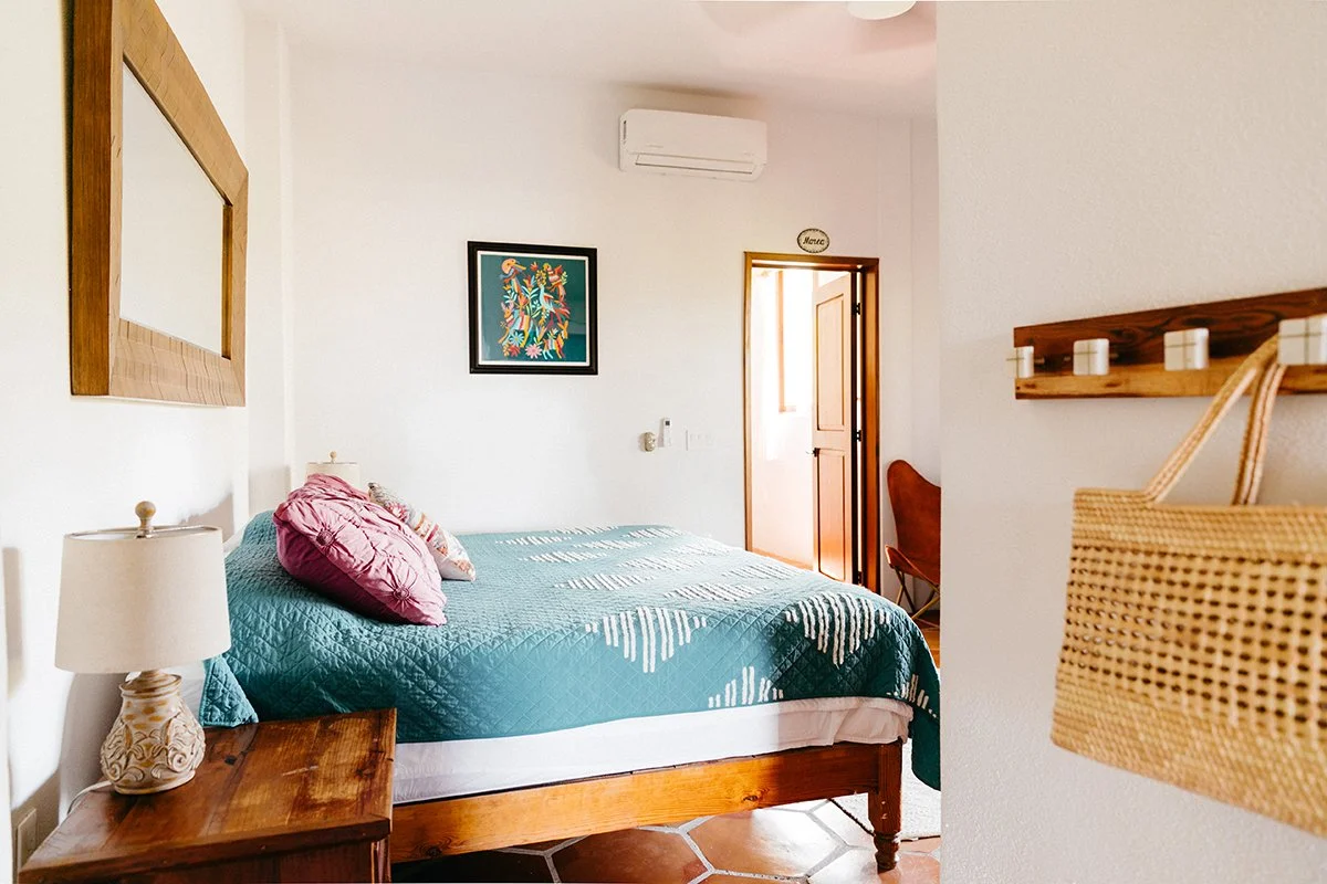 A bedroom with a wooden bed frame and a teal quilt, pink pillows, a nightstand with a lamp, a framed colorful artwork on the white wall, a wooden mirror frame, a small wall decoration reading 'Home,' a doorway leading outside, and a wicker basket on 