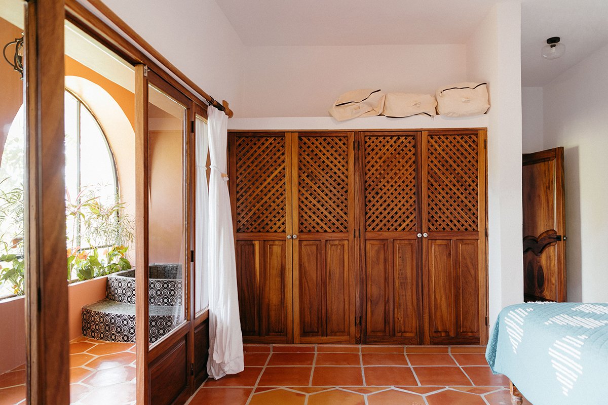 Interior of a room with a wooden closet and a balcony area with potted plants, terracotta tile flooring, and a bed with a light blue blanket.