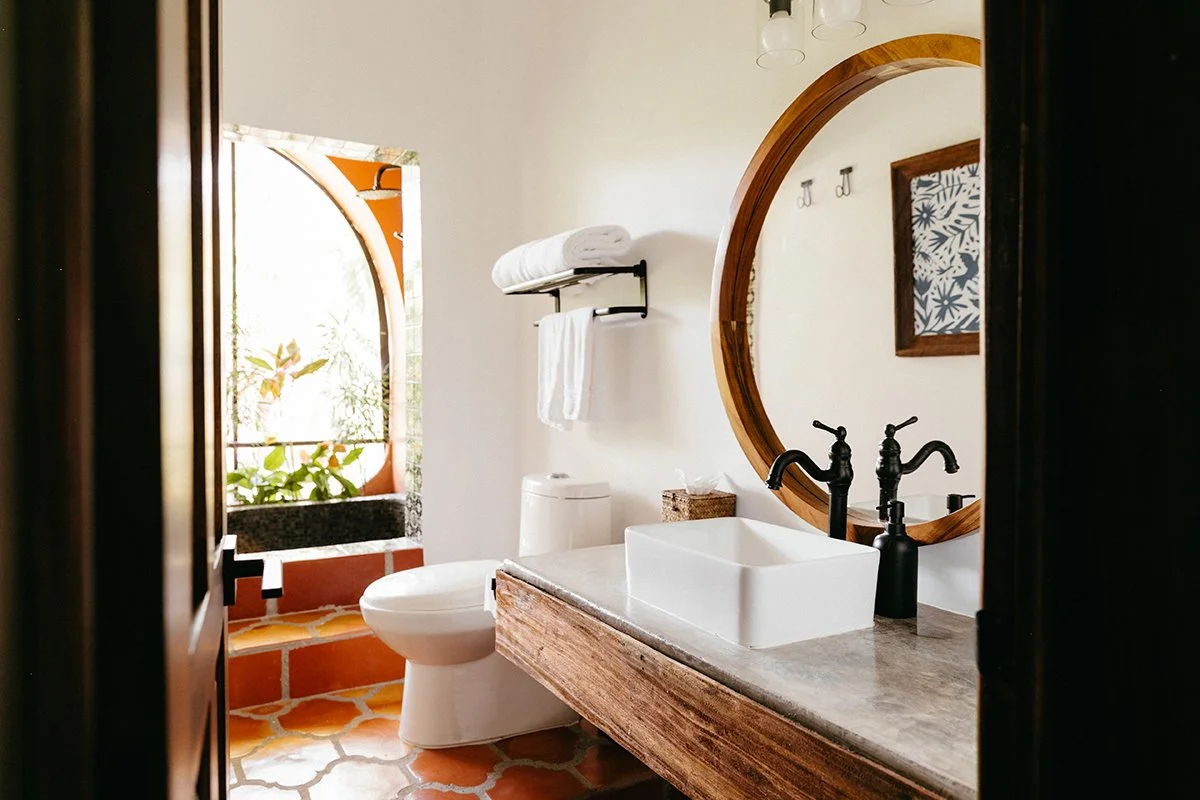 A bathroom with a standing white rectangular sink on a wooden countertop, a black faucet, a round wooden mirror, a toilet, a wall-mounted towel rack with white towels, a window with an arched design and plants outside, terracotta floor tiles, and wal