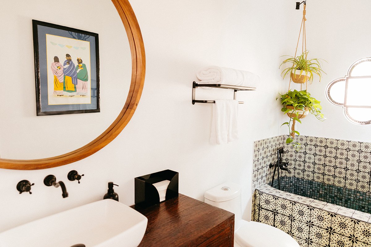 A bathroom featuring a white sink with black fixtures, a large round wooden mirror, a wall-mounted black towel rack with white towels, a decorative bathtub with black and cream patterned tiles, a hanging potted plant, a window, and a framed artwork o