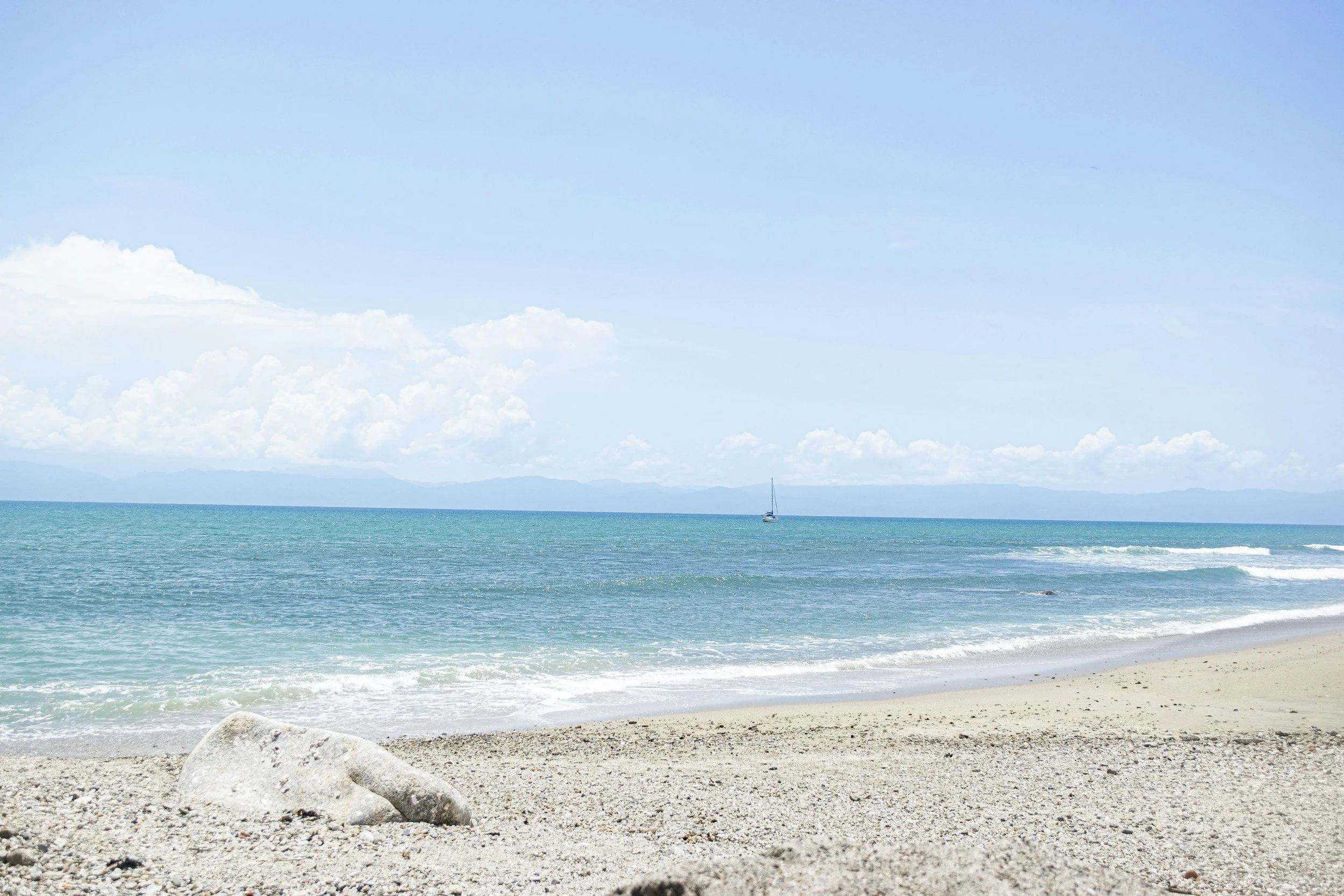 A beach scene with a sandy shoreline, a large rock in the foreground, calm turquoise water, a sailboat in the distance, and a cloudy sky.