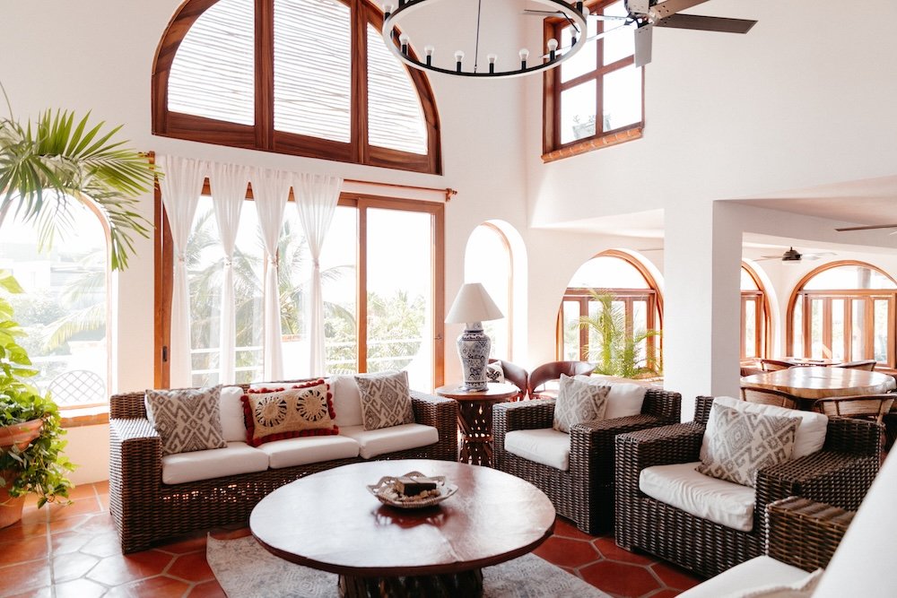 Bright living room with large arched windows, white walls, wicker furniture with white cushions and patterned throw pillows, a round wooden coffee table, a lamp with a decorative blue and white vase base, potted plants, and sheer white curtains, in a