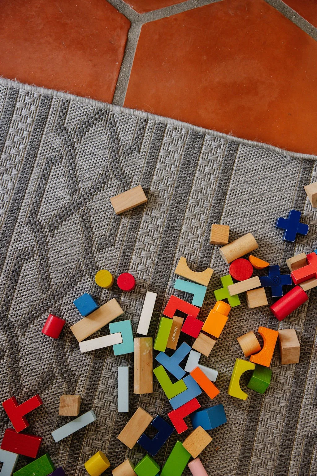 Colorful wooden toy blocks scattered on a gray textured carpet near a tiled floor.