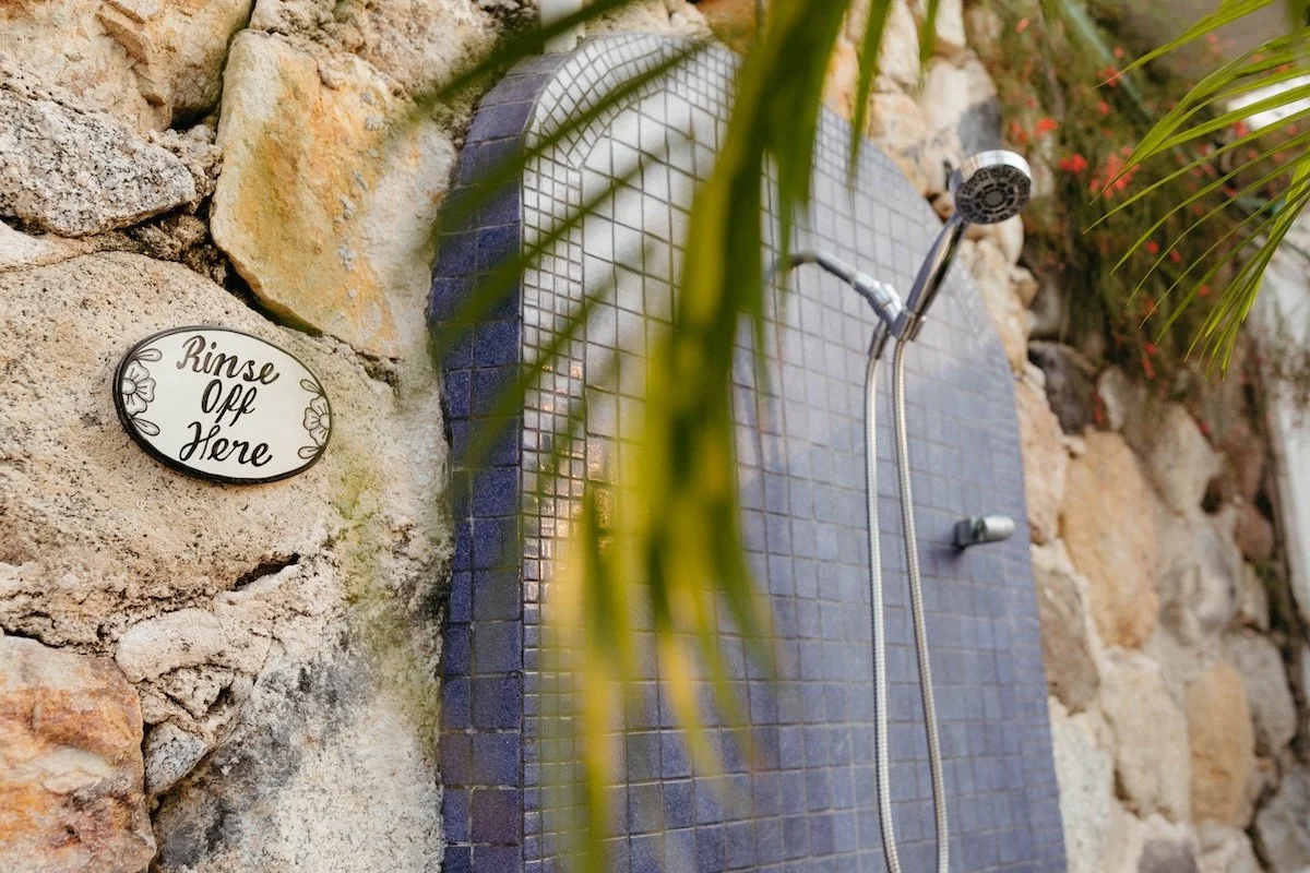 An outdoor shower with a tiled wall, a chrome showerhead, and a sign that says "Rinse Off Here" on a stone wall, surrounded by plants and rocks.