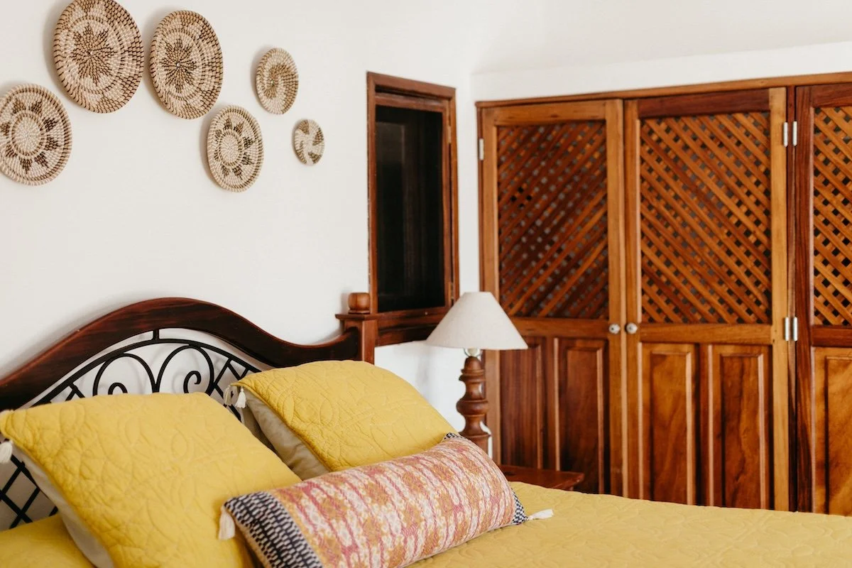 A bedroom with a wooden bed frame and yellow bedding, including yellow pillows and a decorative pillow. There is a wooden nightstand with a lamp next to the bed, a window, and a large wooden wardrobe. Decorative woven baskets hang on the white wall a