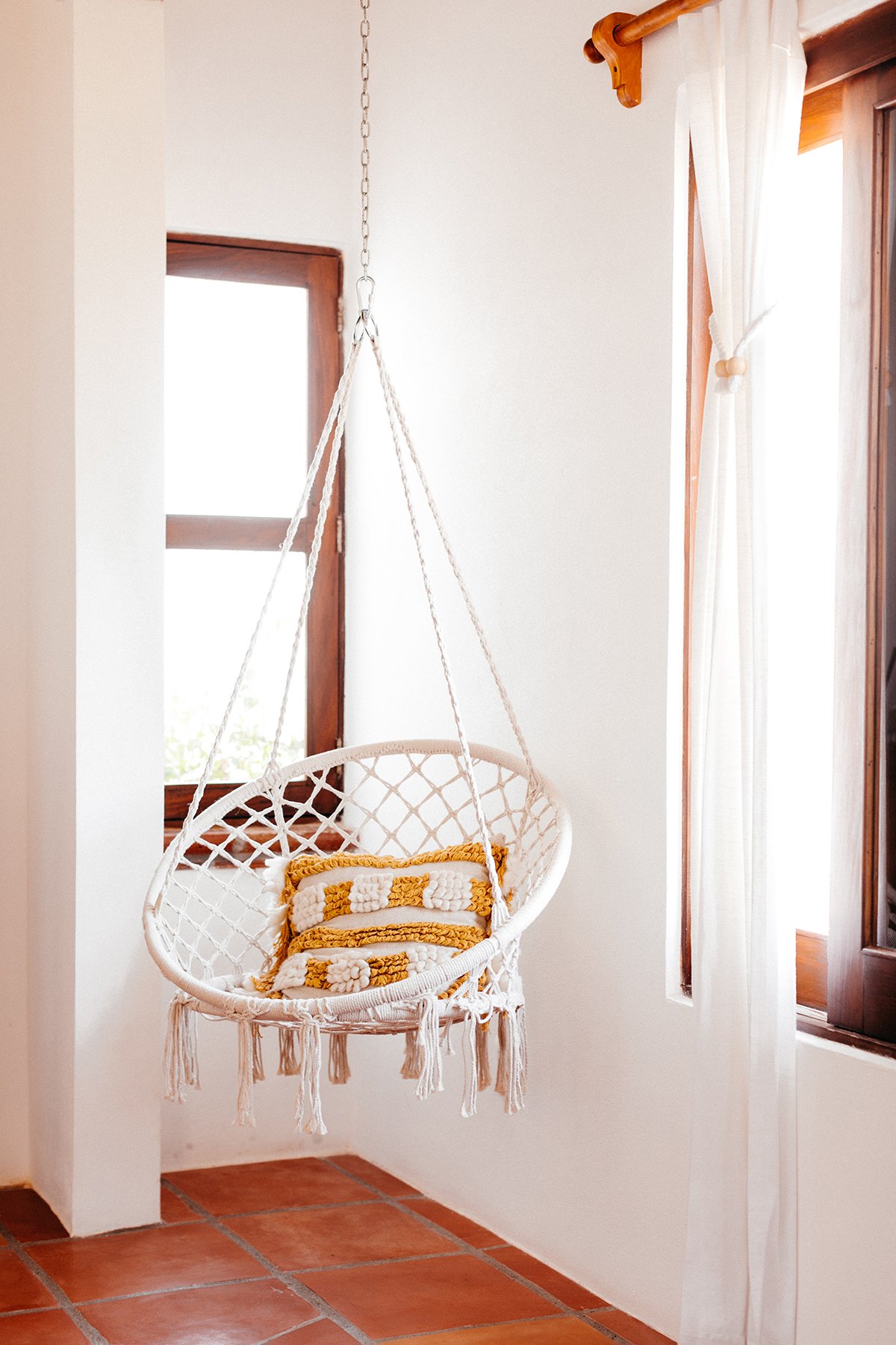 A cozy indoor space featuring a white hanging chair with a yellow and white textured pillow, located next to a window with white curtains and wooden trim, with terracotta flooring.