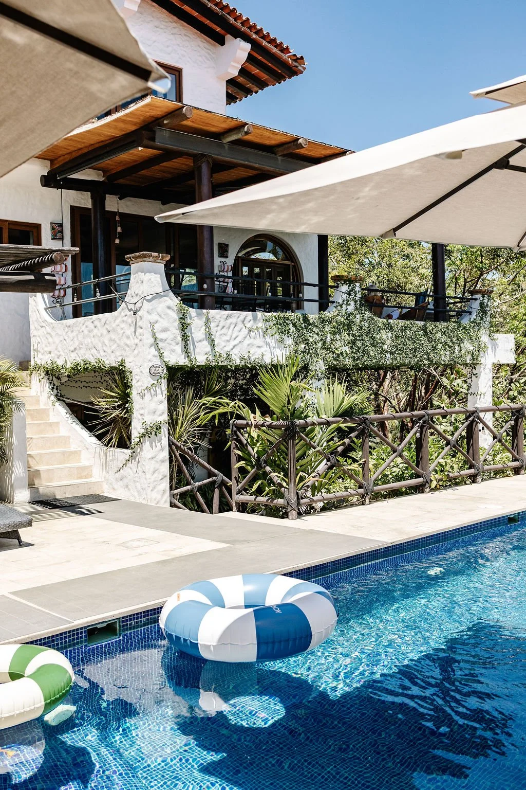 A swimming pool with float rings, surrounded by a patio and lush greenery, with a white building featuring a balcony and large umbrellas providing shade.