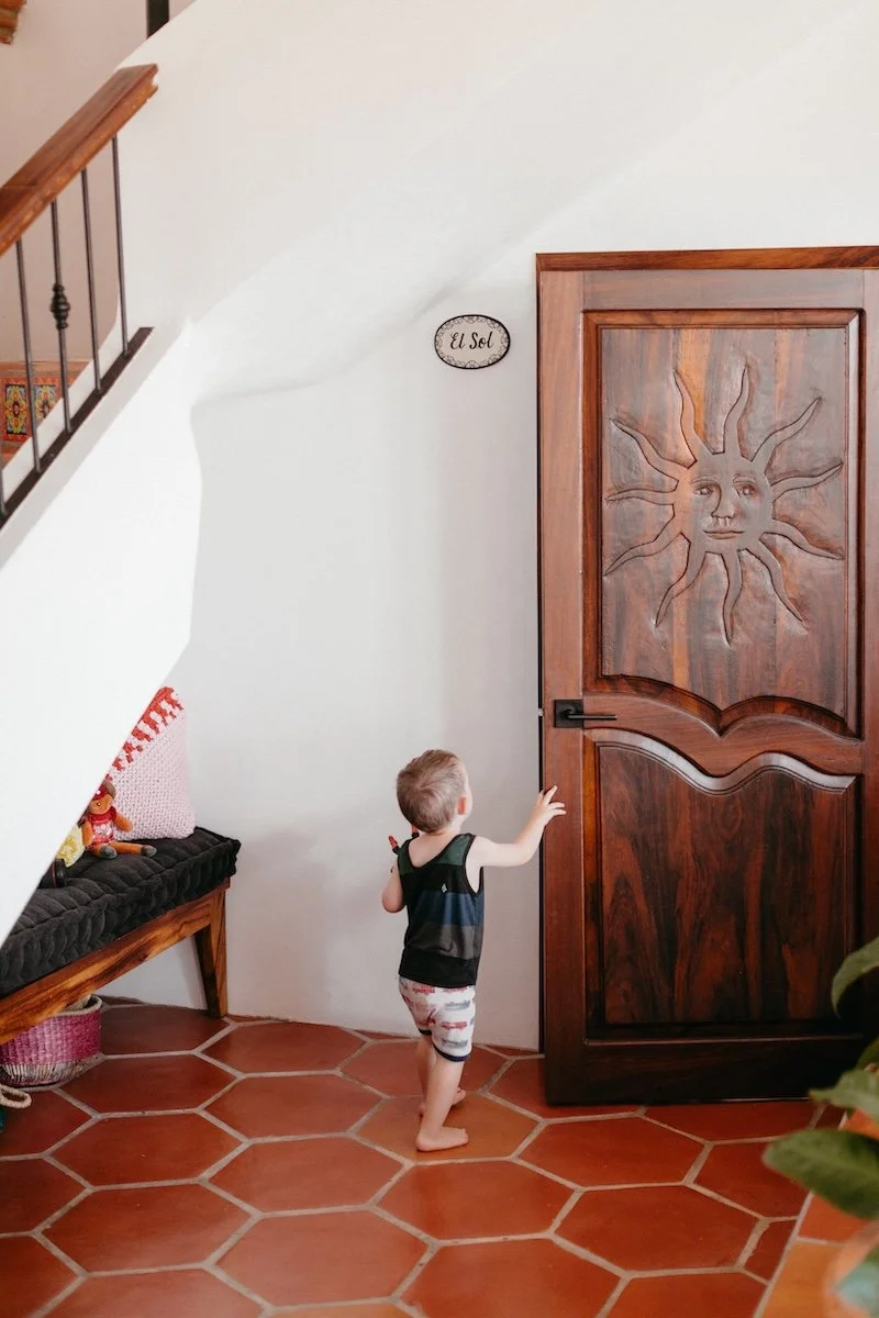 A young boy wearing a tank top and shorts is standing in front of a wooden door with a sun face carved into it. The scene appears to be inside a home with tiled floor and a bench with plush cushion and decorative items nearby.