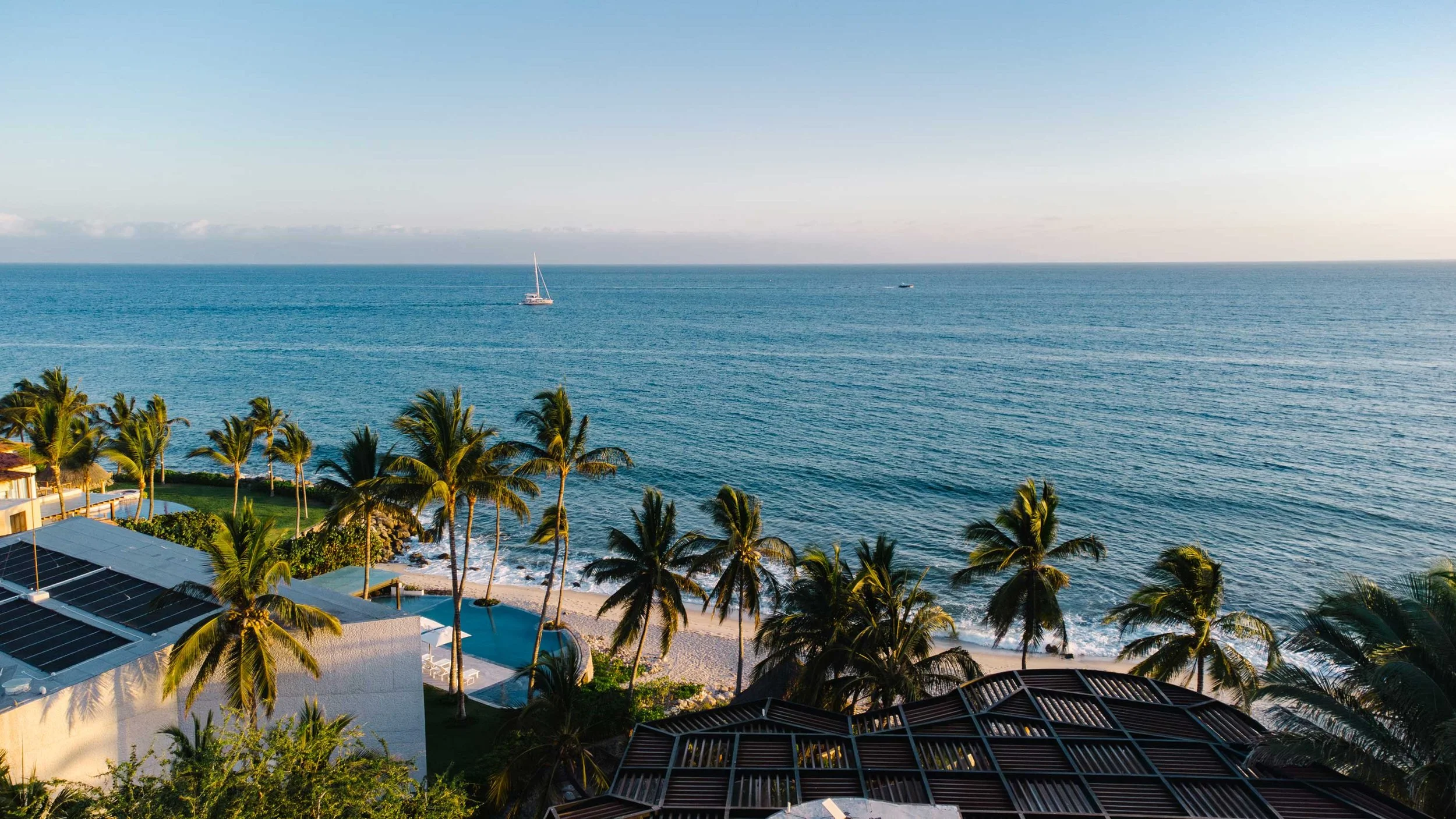 A tropical beach view with palm trees, buildings, and a swimming pool in the foreground, the ocean with a sailboat and a small boat in the distance, and clear blue sky.