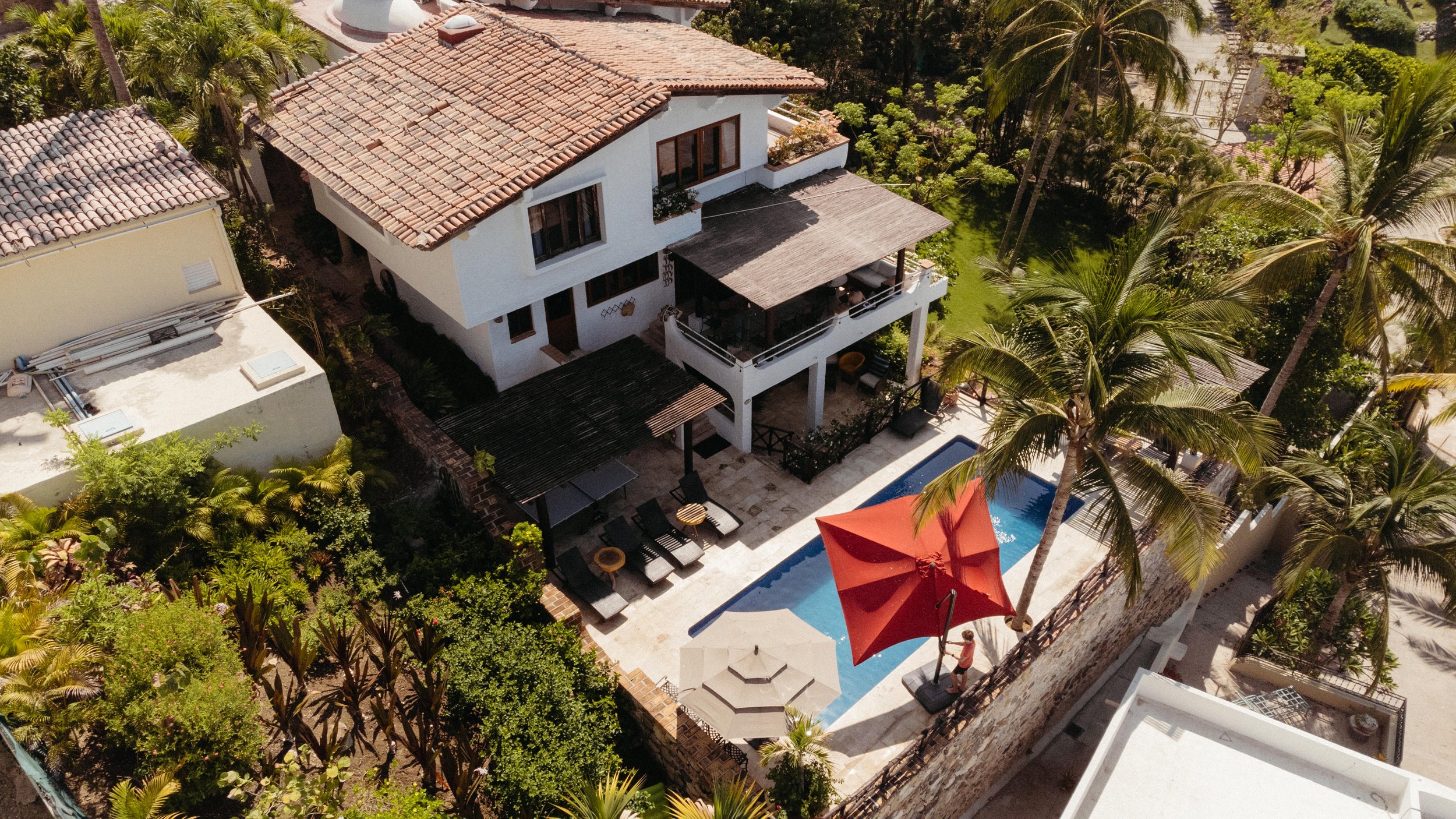An aerial view of a beautiful house with a terracotta tiled roof, surrounded by lush tropical trees. The backyard features a swimming pool with a large red umbrella, lounge chairs, and a covered patio area.