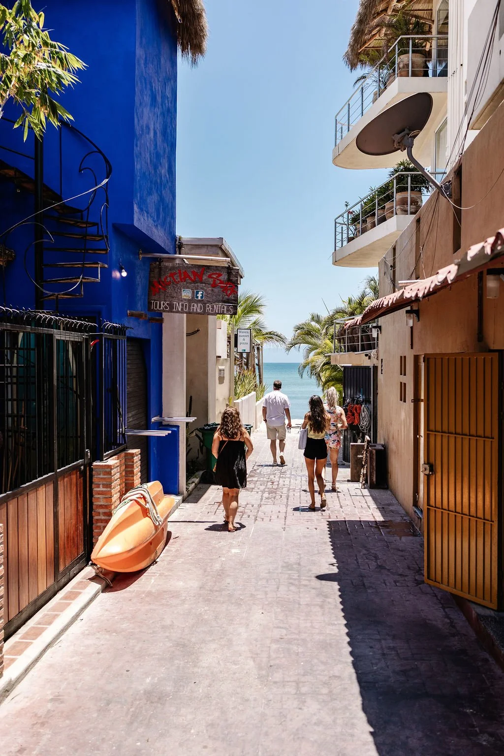 People walking down a narrow alley toward the beach, with palm trees and ocean visible in the background. Colorful buildings with balconies and a sign for tours and rentals are on either side.