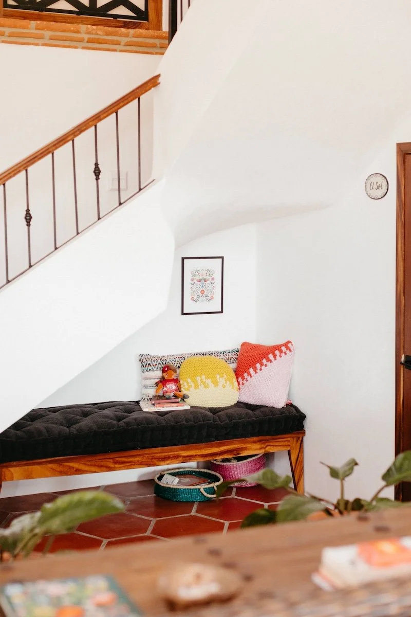 Interior of a cozy home corner with a wooden bench, black cushion, and colorful pillows, a small doll, and books on the bench. A framed artwork hangs on the white wall above. A staircase with a wooden handrail and black iron balusters is on the left.