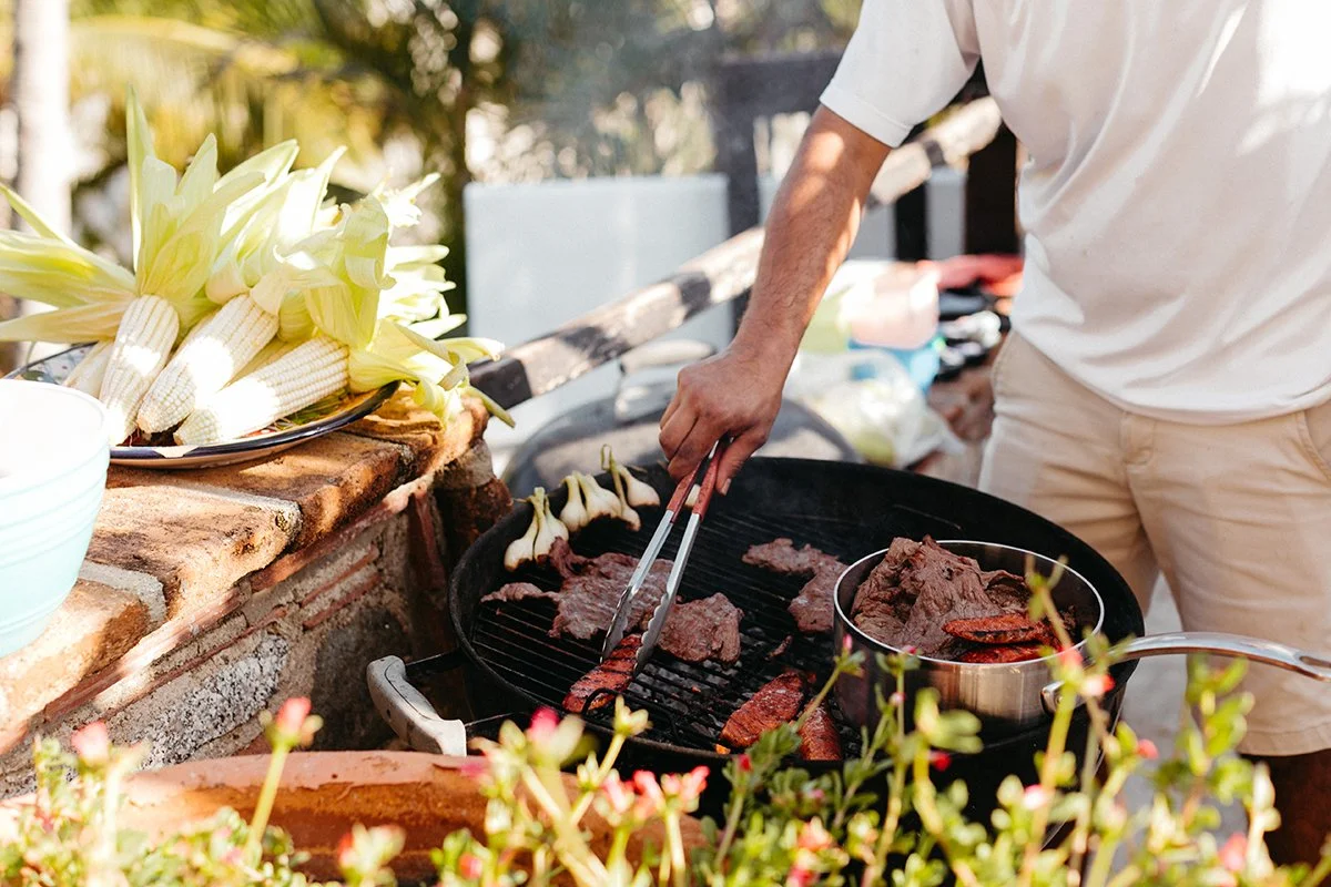Person cooking steaks and vegetables on an outdoor grill surrounded by fresh produce and flowers.