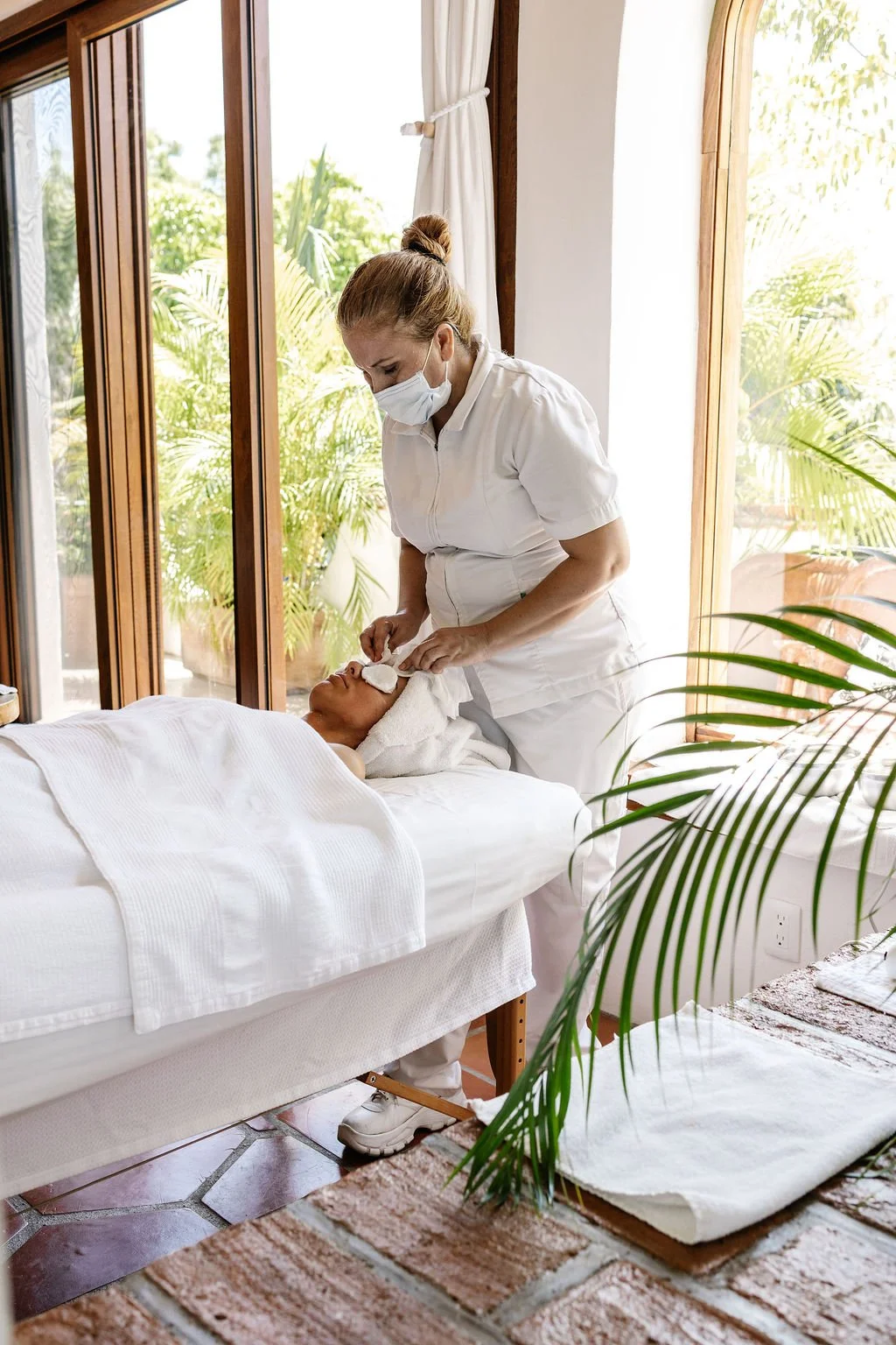 A nurse in a white uniform and face mask provides facial treatment to a woman lying in bed in a bright, tropical-style room with large windows and greenery outside.