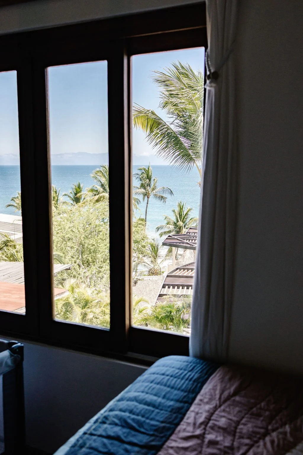 View of palm trees, ocean, and sky seen through a large window from inside a room with a bed and curtains.