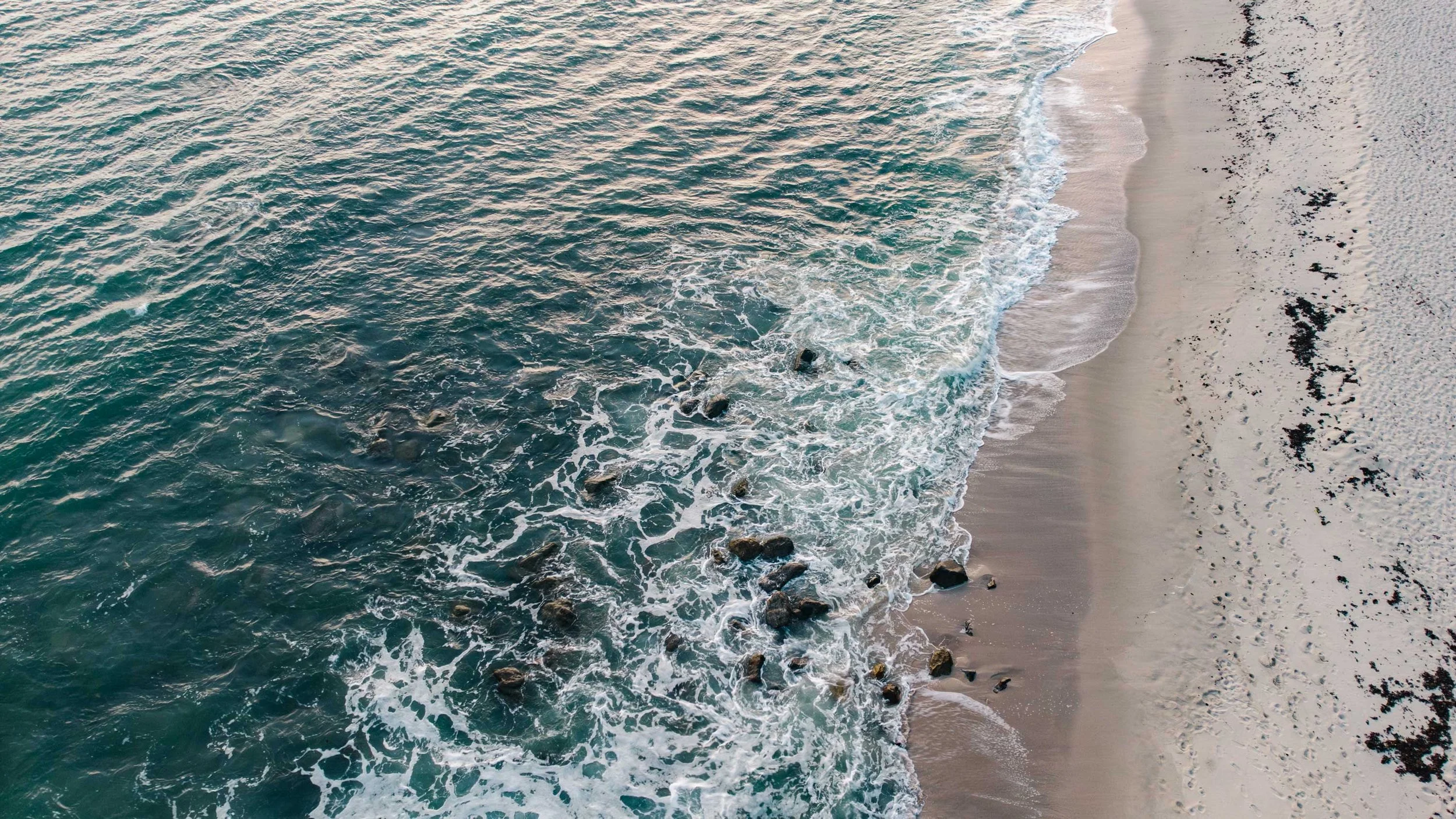 Aerial view of the ocean waves crashing onto a sandy beach with rocks and seaweed.