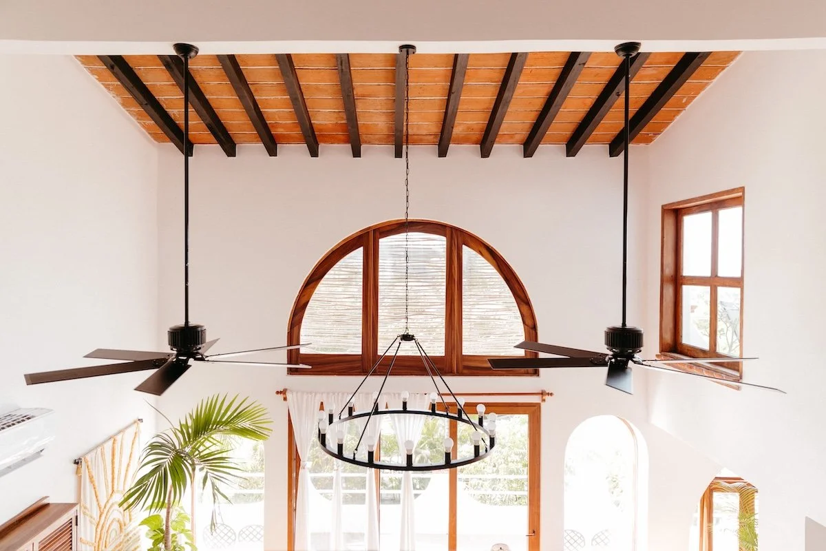 Interior view of a bright living room with high vaulted ceiling featuring exposed wooden beams, large arched window with white curtains, rectangular window, ceiling fans, a large circular chandelier, and potted plants.
