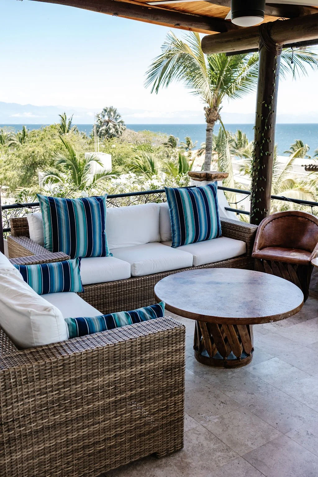 Outdoor patio with wicker furniture and striped cushions, overlooking tropical trees and the ocean in the background.