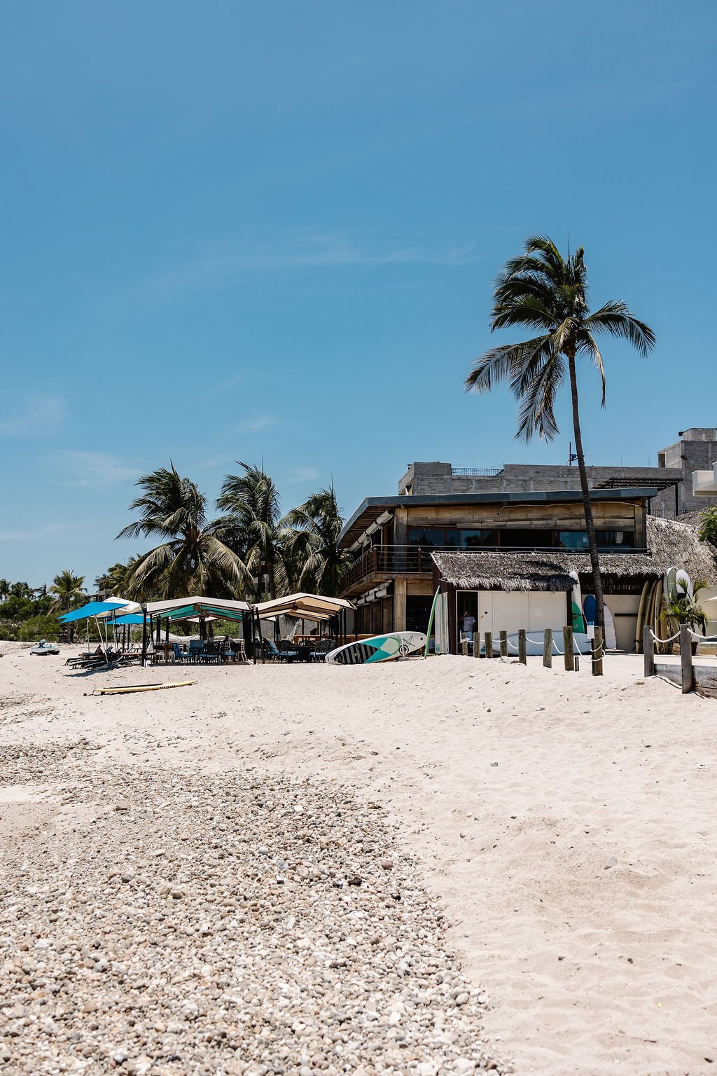 Beach scene with white sand, palm trees, beach umbrellas, and a modern building in the background under a clear blue sky.