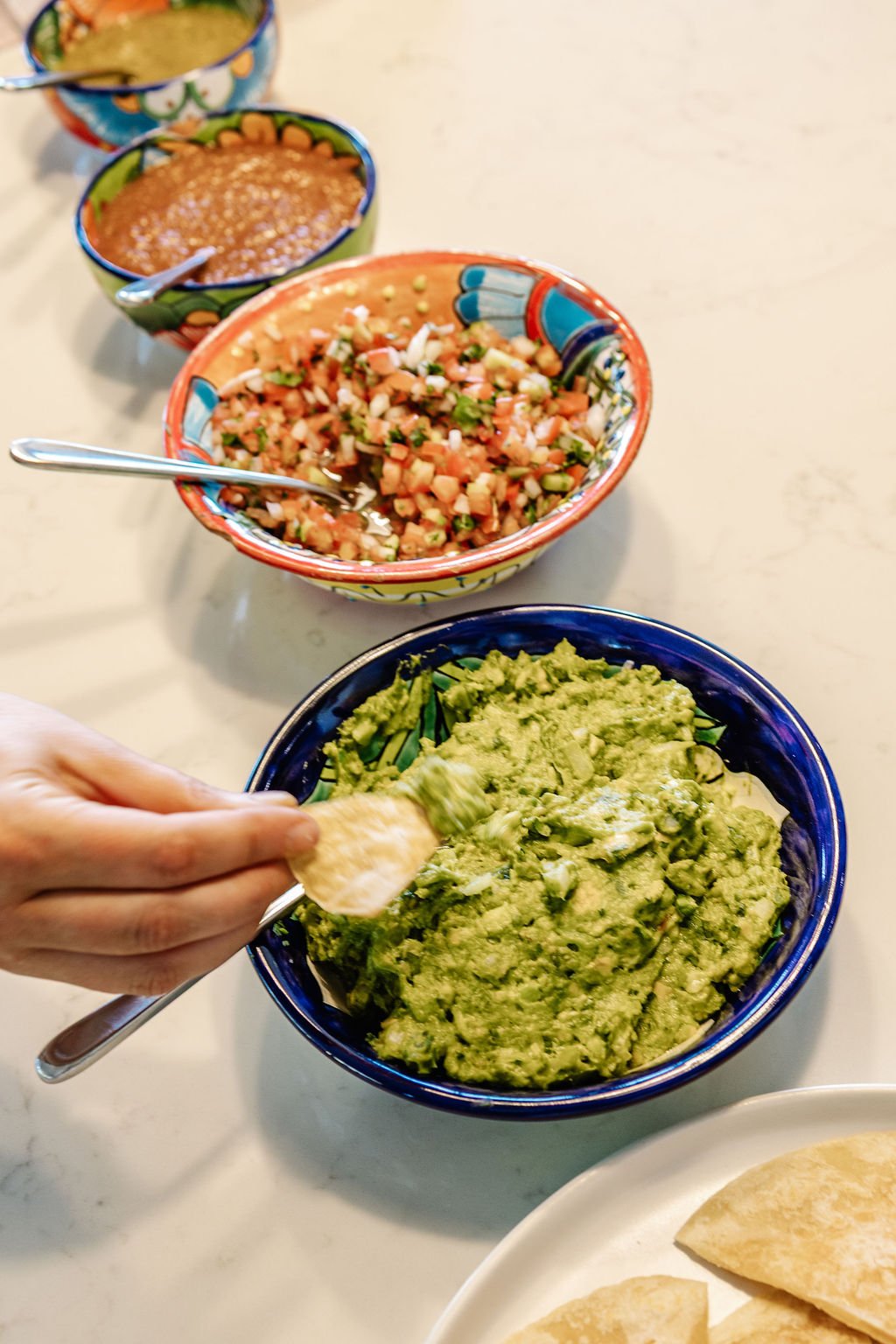 Bowl of guacamole being served with a chip, surrounded by bowls of pico de gallo, beans, and salsa on a white table.