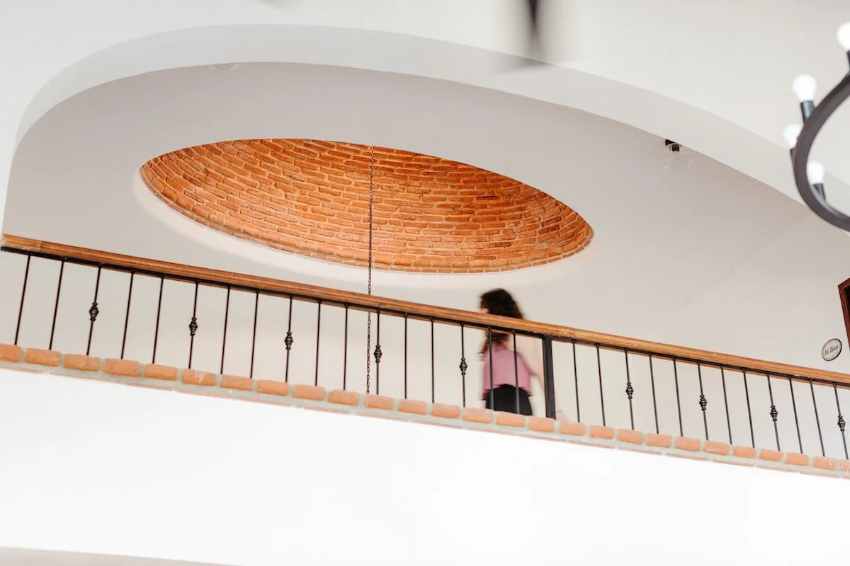 A person walking on an upper level balcony with a black metal railing and a wooden handrail, beneath an oval-shaped opening in the ceiling with exposed red brick interior.